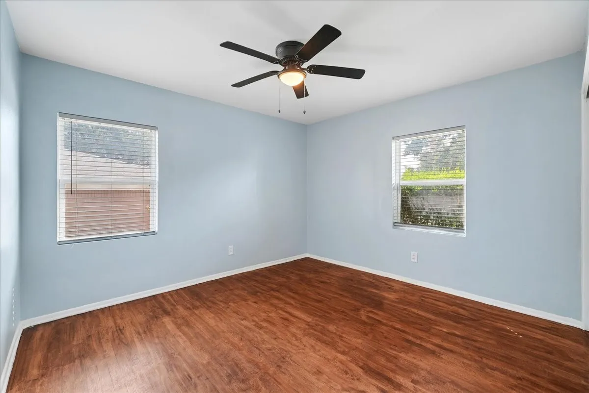 Room with wood-finish flooring, light blue walls, two windows with horizontal blinds, and a ceiling fan with integrated lighting