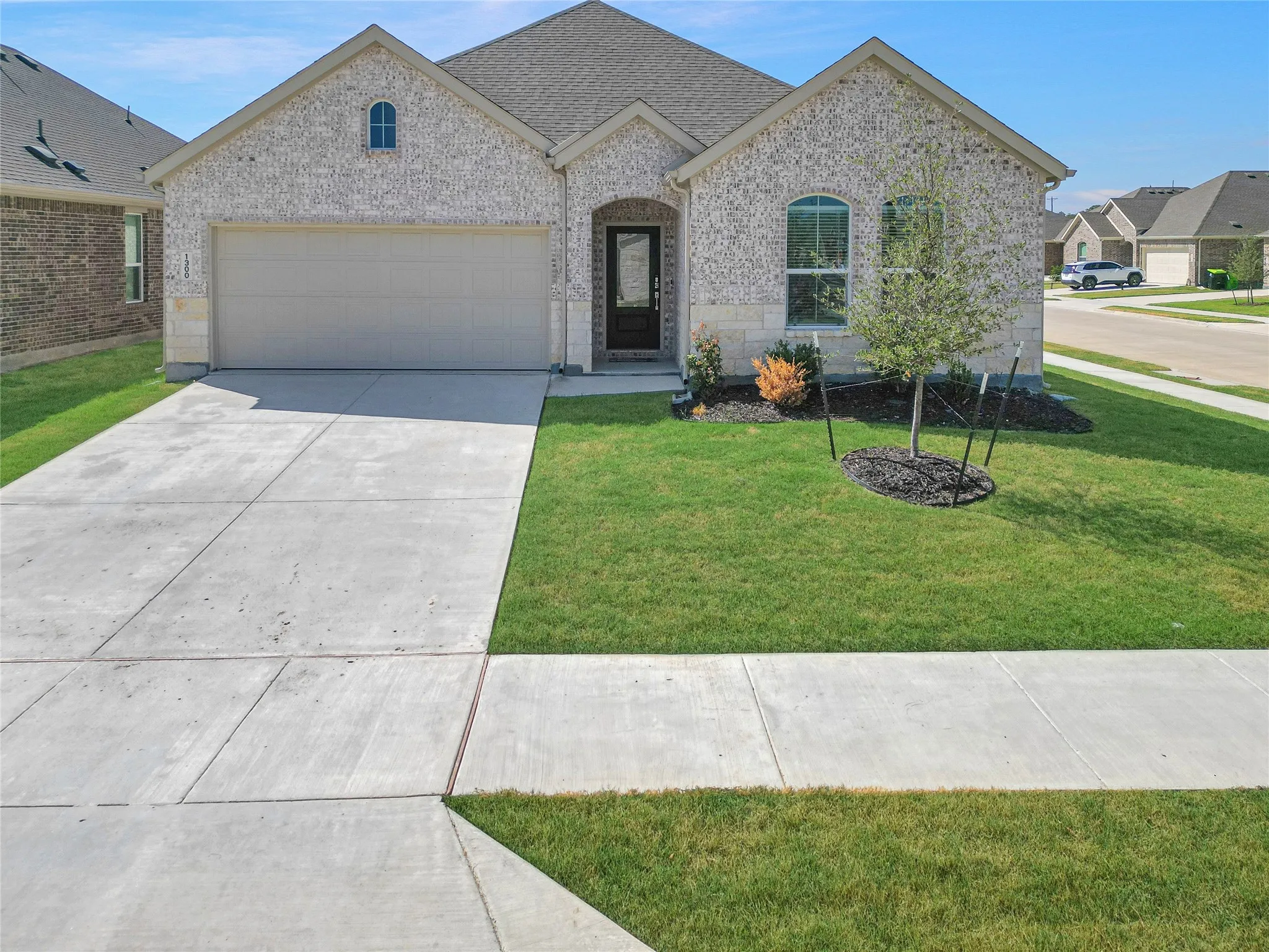 Light-colored brick exterior with stone accents, an attached garage, a dark-colored front door, and a manicured lawn with a young tree