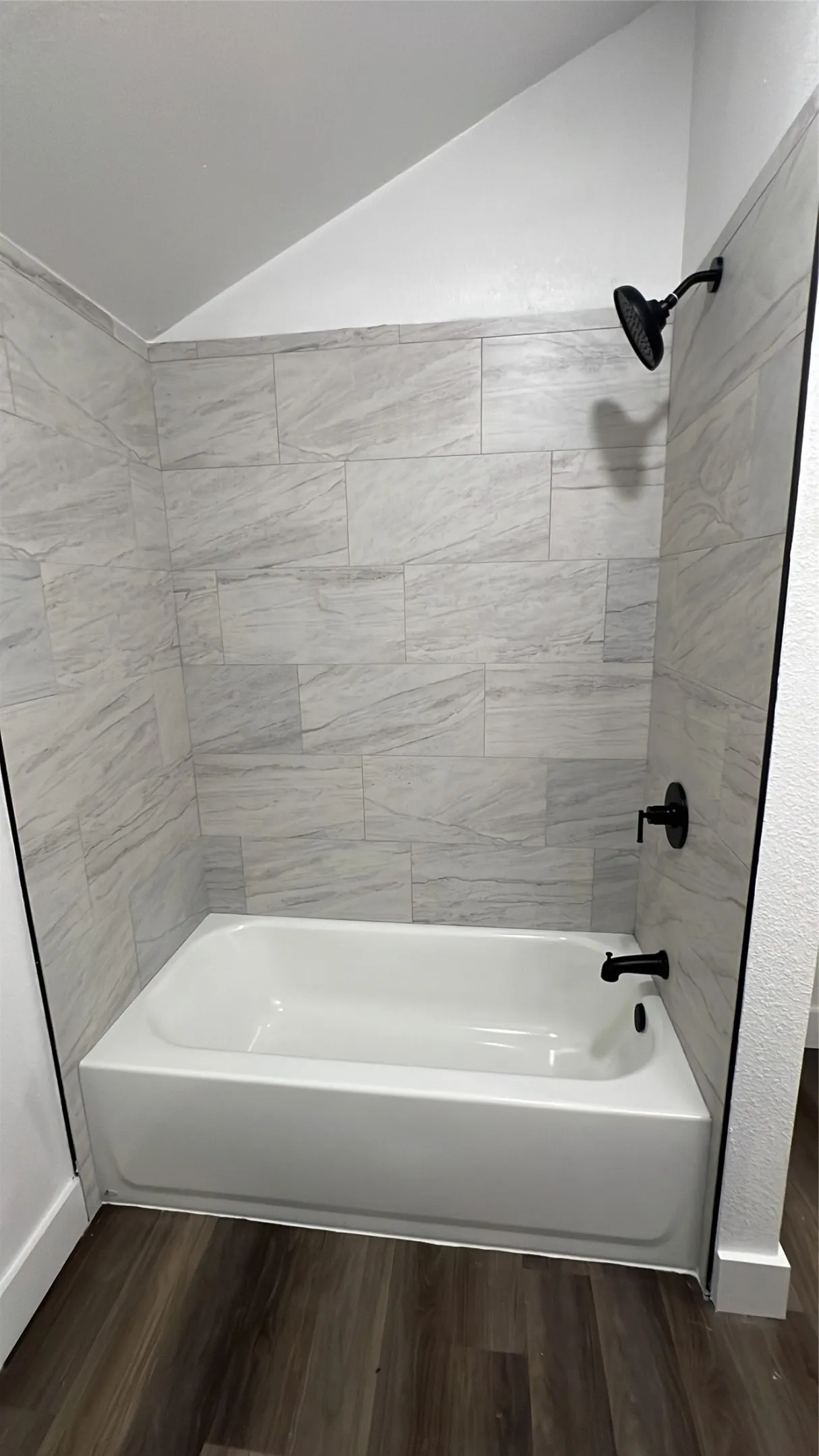 White bathtub with matte black fixtures, surrounded by light-toned tiled walls and wood-finish flooring