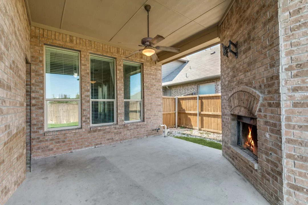 Covered patio featuring a brick exterior, outdoor fireplace, ceiling fan with integrated lighting, and concrete flooring.   Notice in the back there's a separate gas line for barbecue and a TV mount over the fireplace.