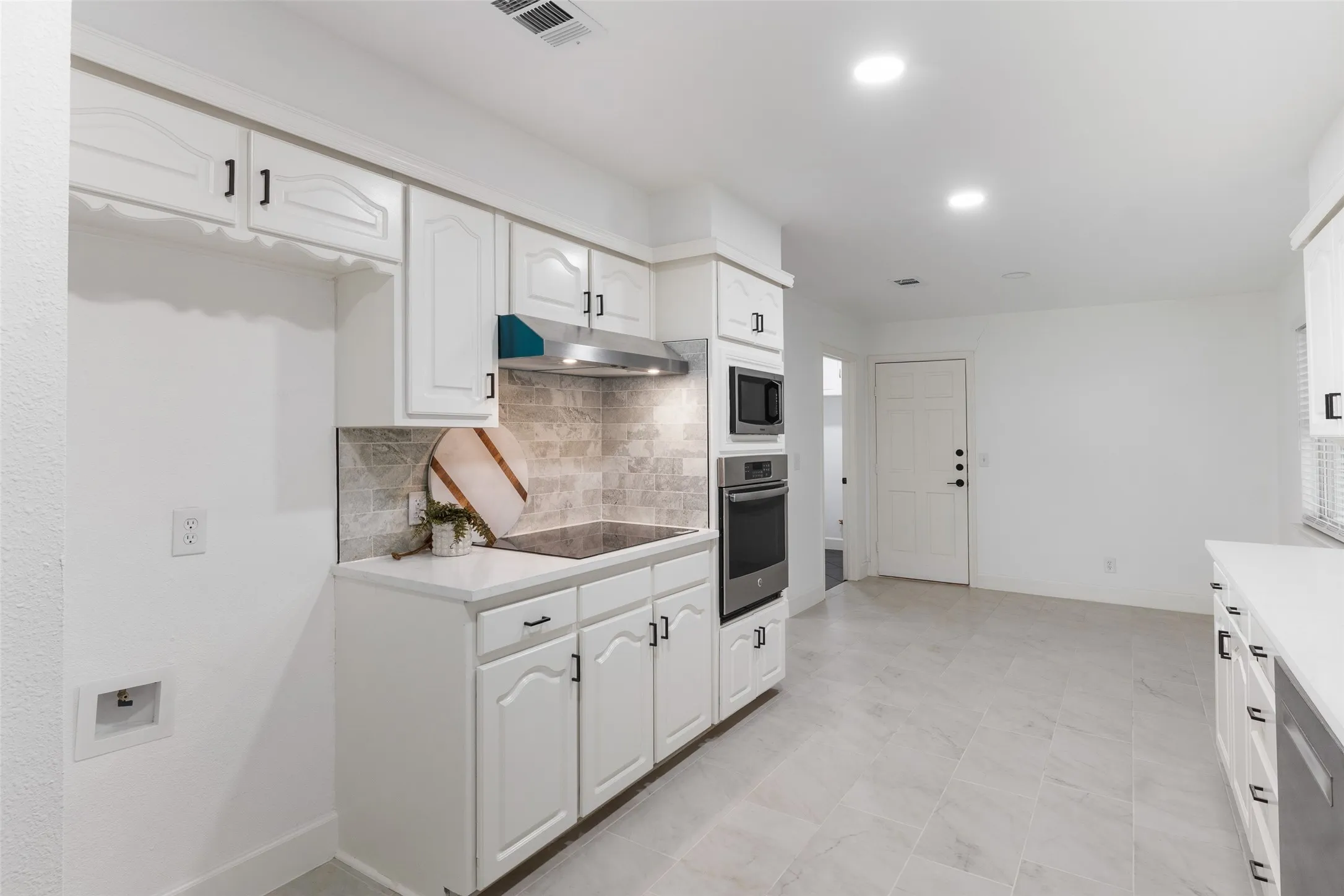 White cabinetry with black hardware, stainless steel built-in oven and microwave, stone-finish backsplash, recessed lighting, and large format tile flooring