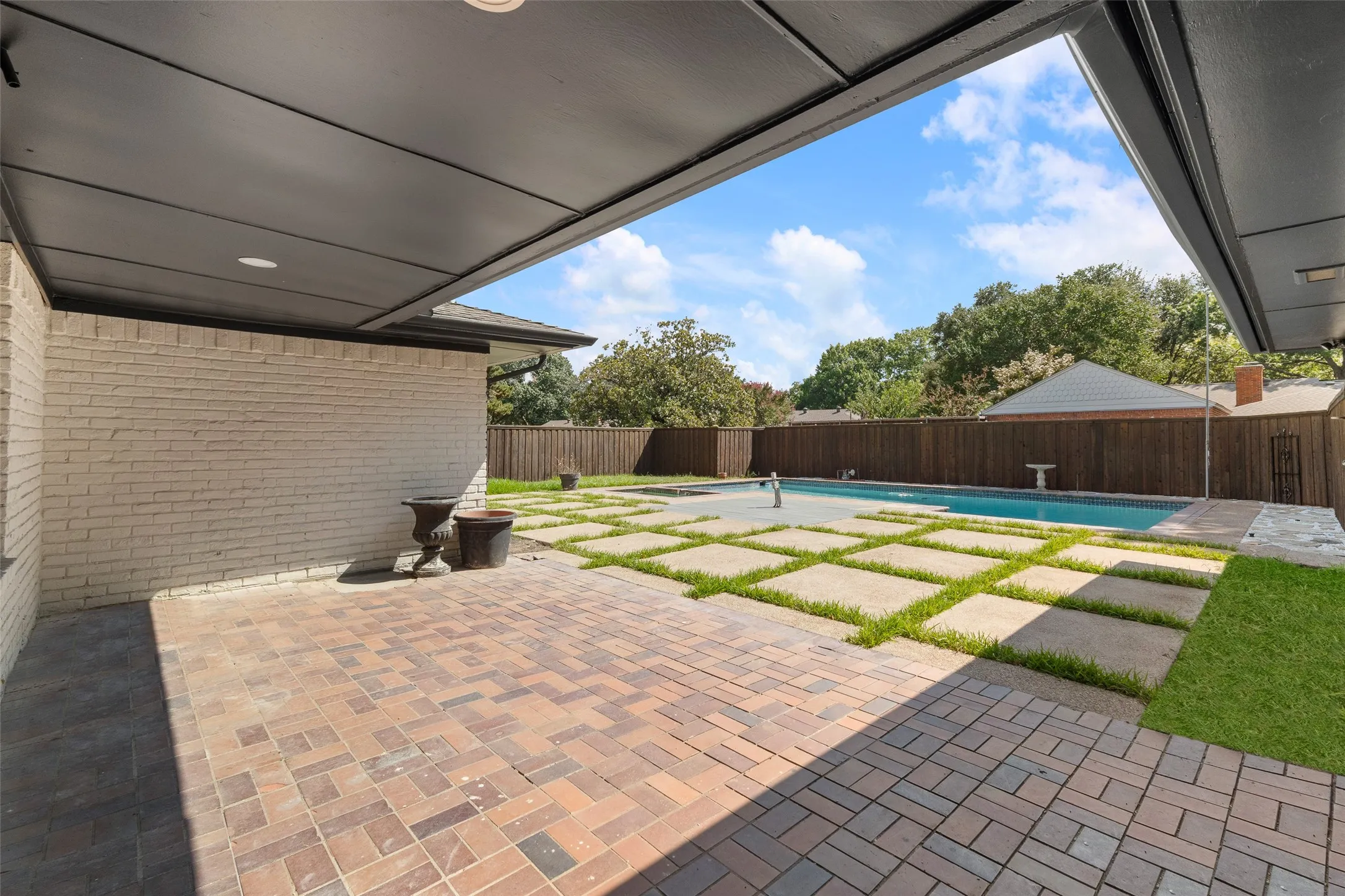 Covered outdoor patio with paver flooring, white brick exterior, mature privacy fencing, concrete stepping stones, and a rectangular swimming pool