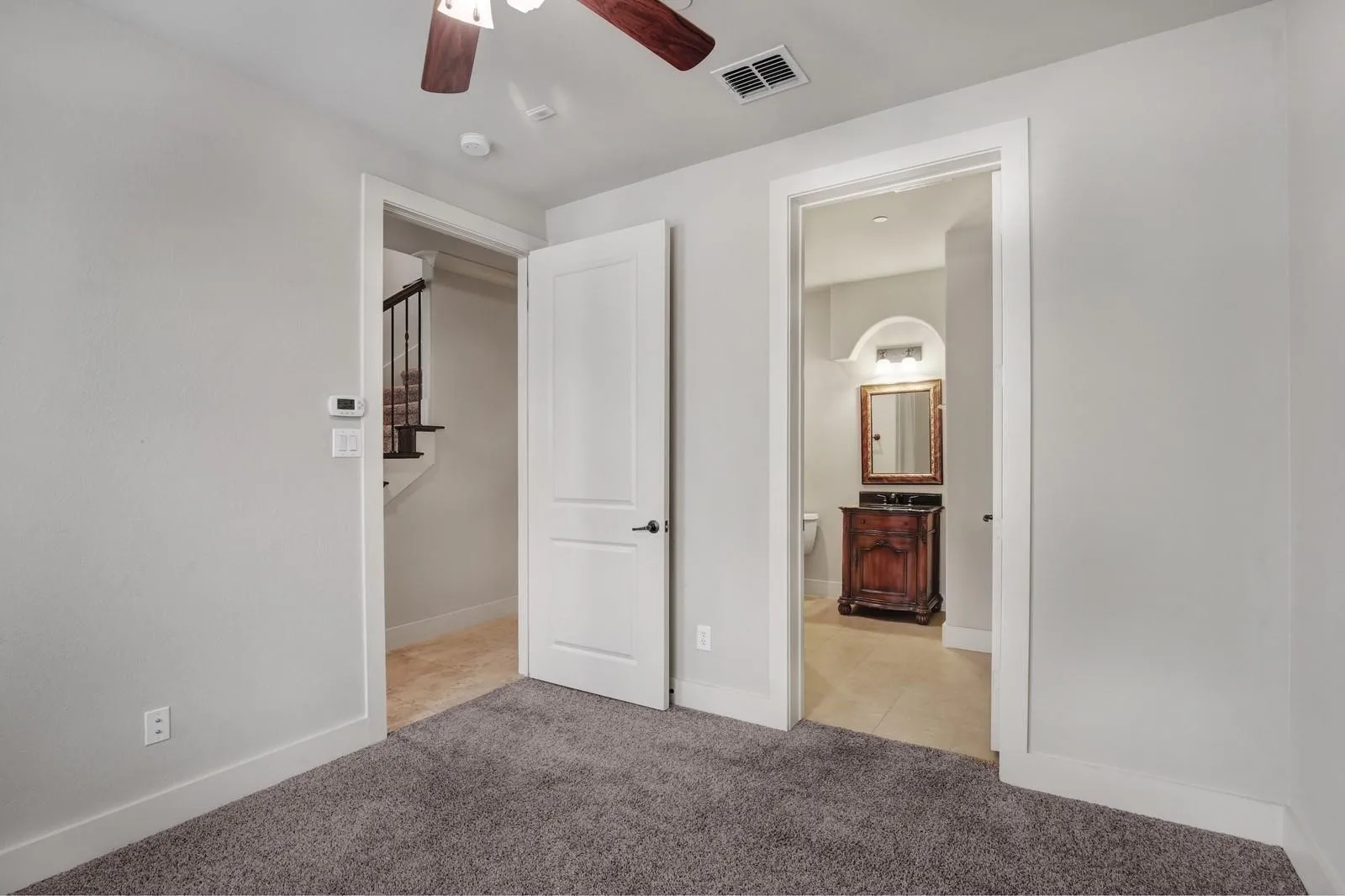 Carpeted room with a ceiling fan, white paneled door, and access to a bathroom featuring a wood-finish vanity and tiled flooring