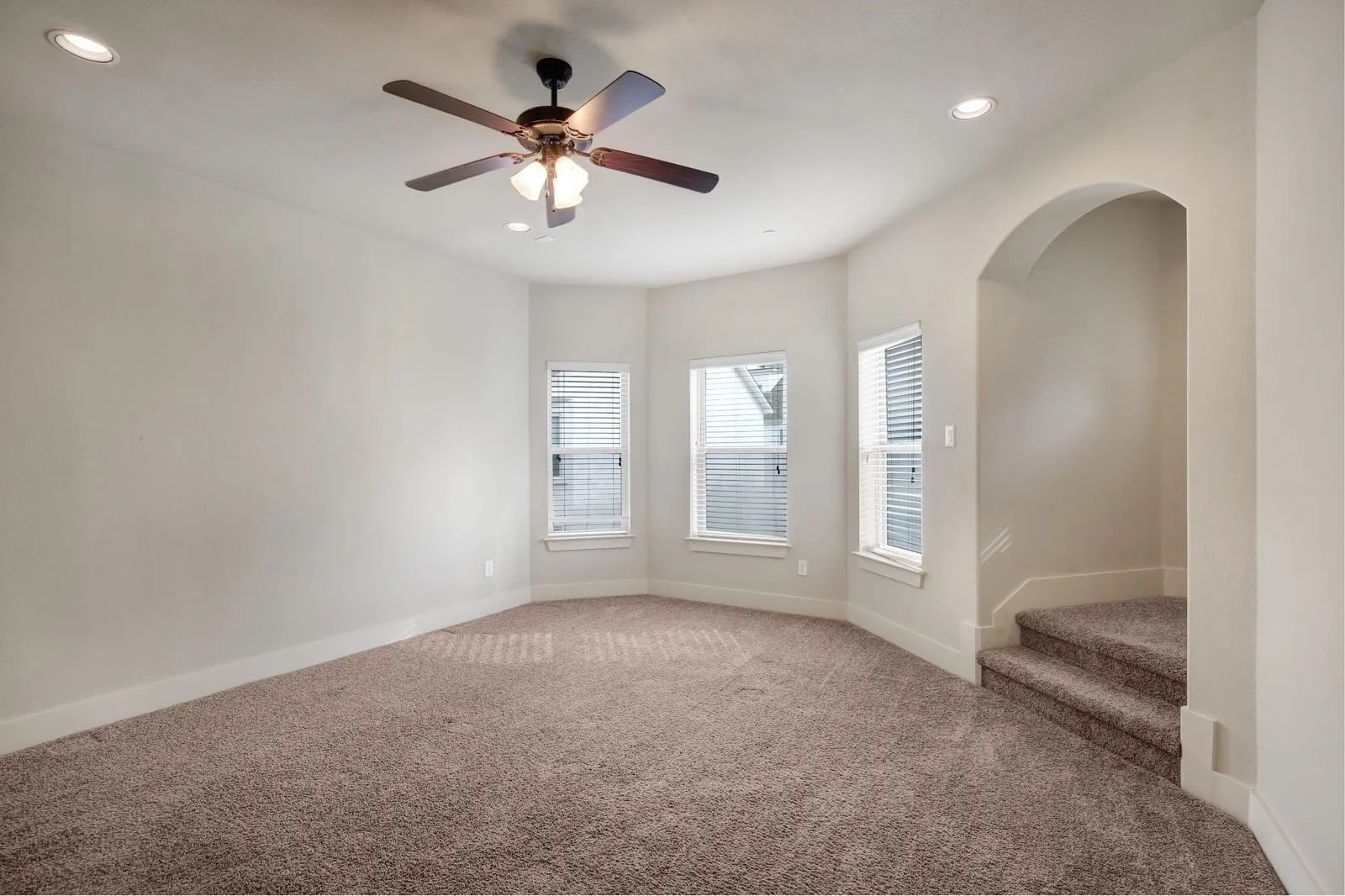 Carpeted room featuring a ceiling fan, recessed lighting, multiple windows with blinds, and an arched entryway leading to carpeted stairs