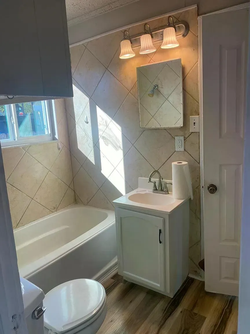 Bathtub with tiled surround, single vanity with integrated sink, wall-mounted light fixture, and wood-finish flooring