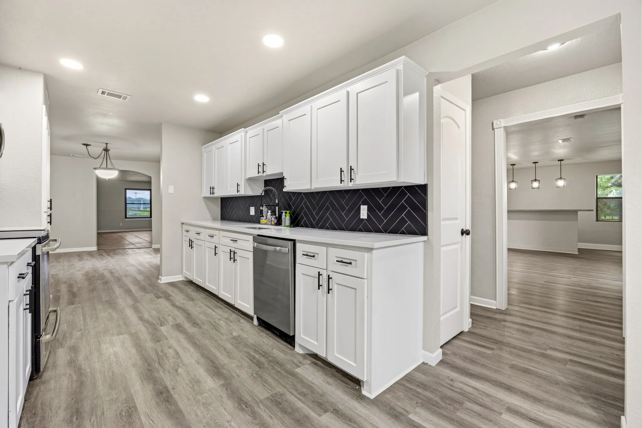 Kitchen featuring white shaker cabinetry, stainless steel dishwasher, black chevron tile backsplash, light-toned countertops, and wood-finish flooring