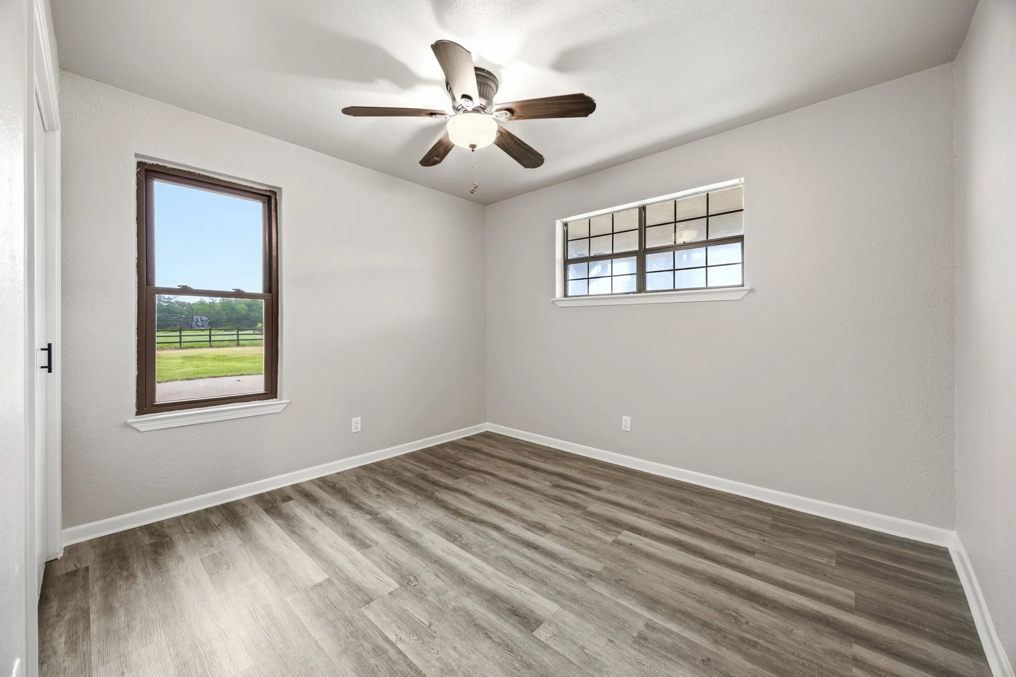 Wood-finish flooring, ceiling fan with integrated lighting, two windows with dark trim, and light-toned walls