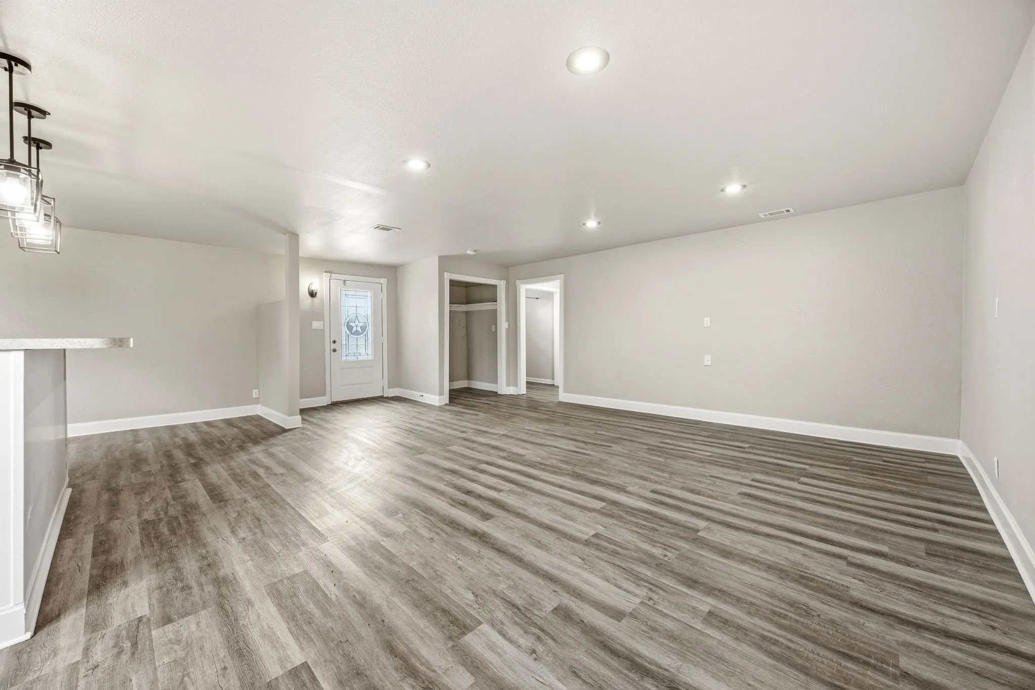 Spacious living area featuring wood-finish flooring, recessed lighting, a white entry door with decorative glass, and a built-in breakfast bar with a solid surface countertop
