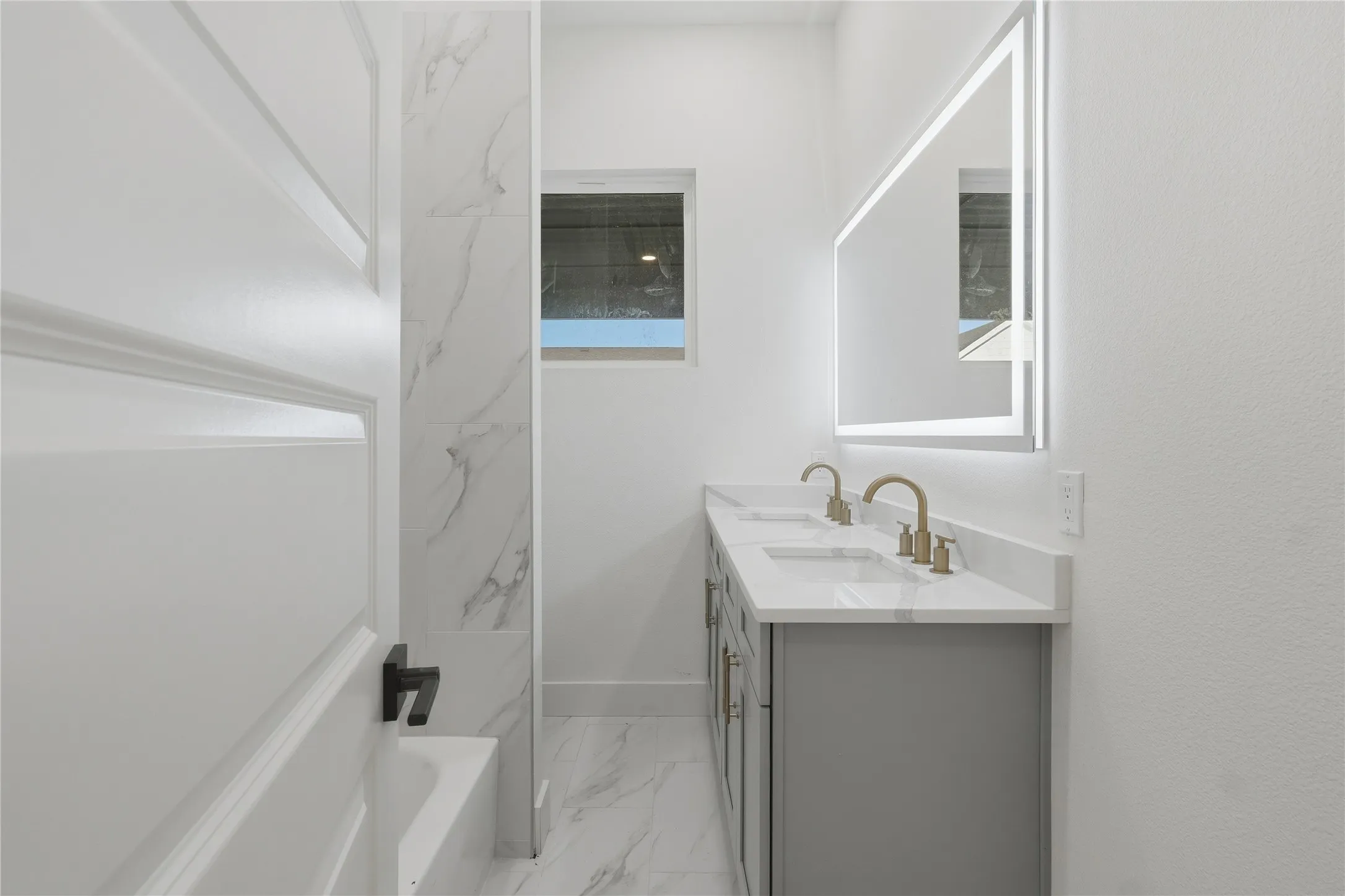 Dual vanity with light gray cabinetry and gold-tone fixtures, under-mount sinks, and a backlit mirror