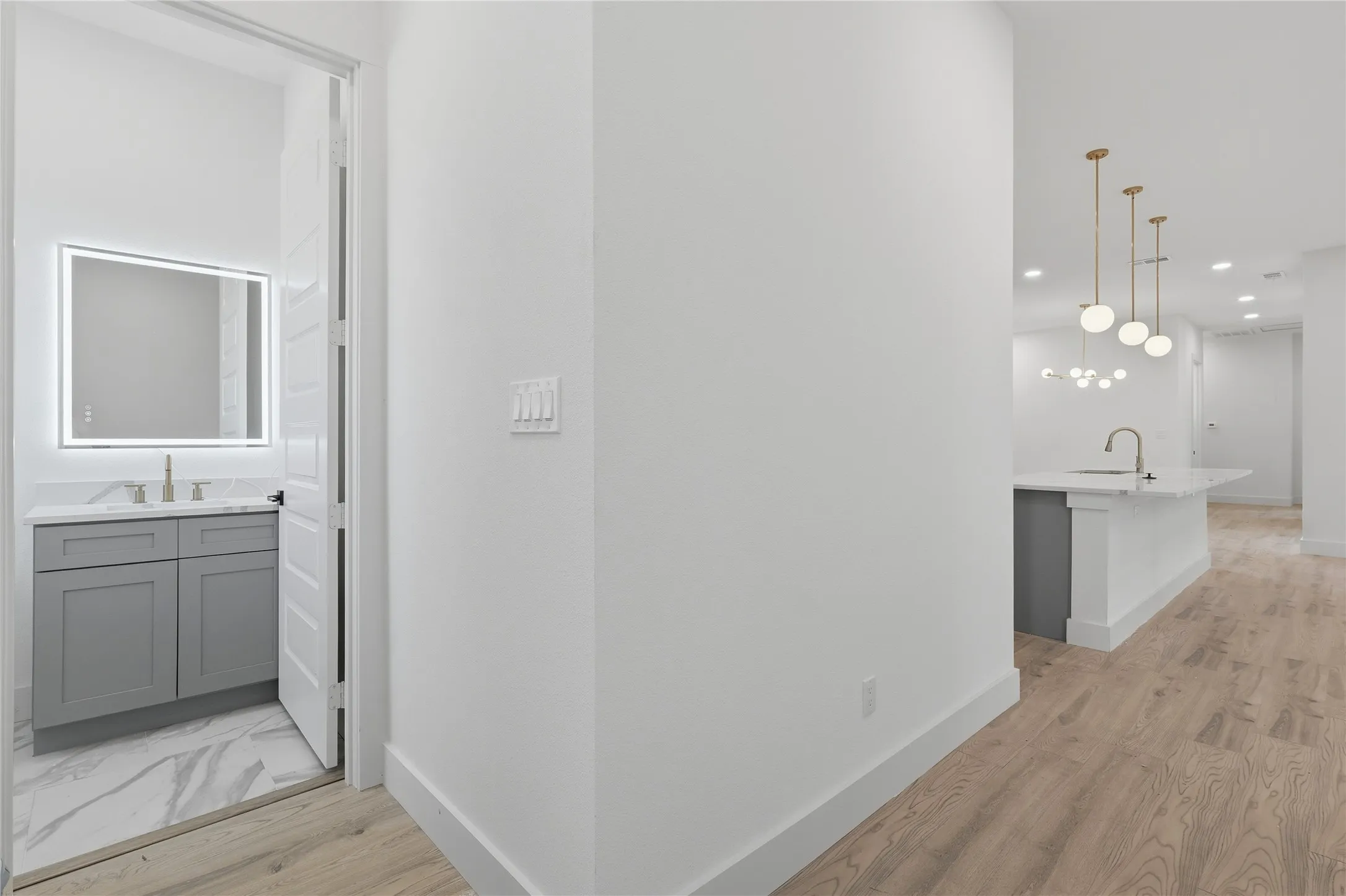 Wood-finish flooring throughout, modern kitchen island with white countertop, contemporary pendant lighting, recessed lighting, and a bathroom with a backlit mirror and gray vanity