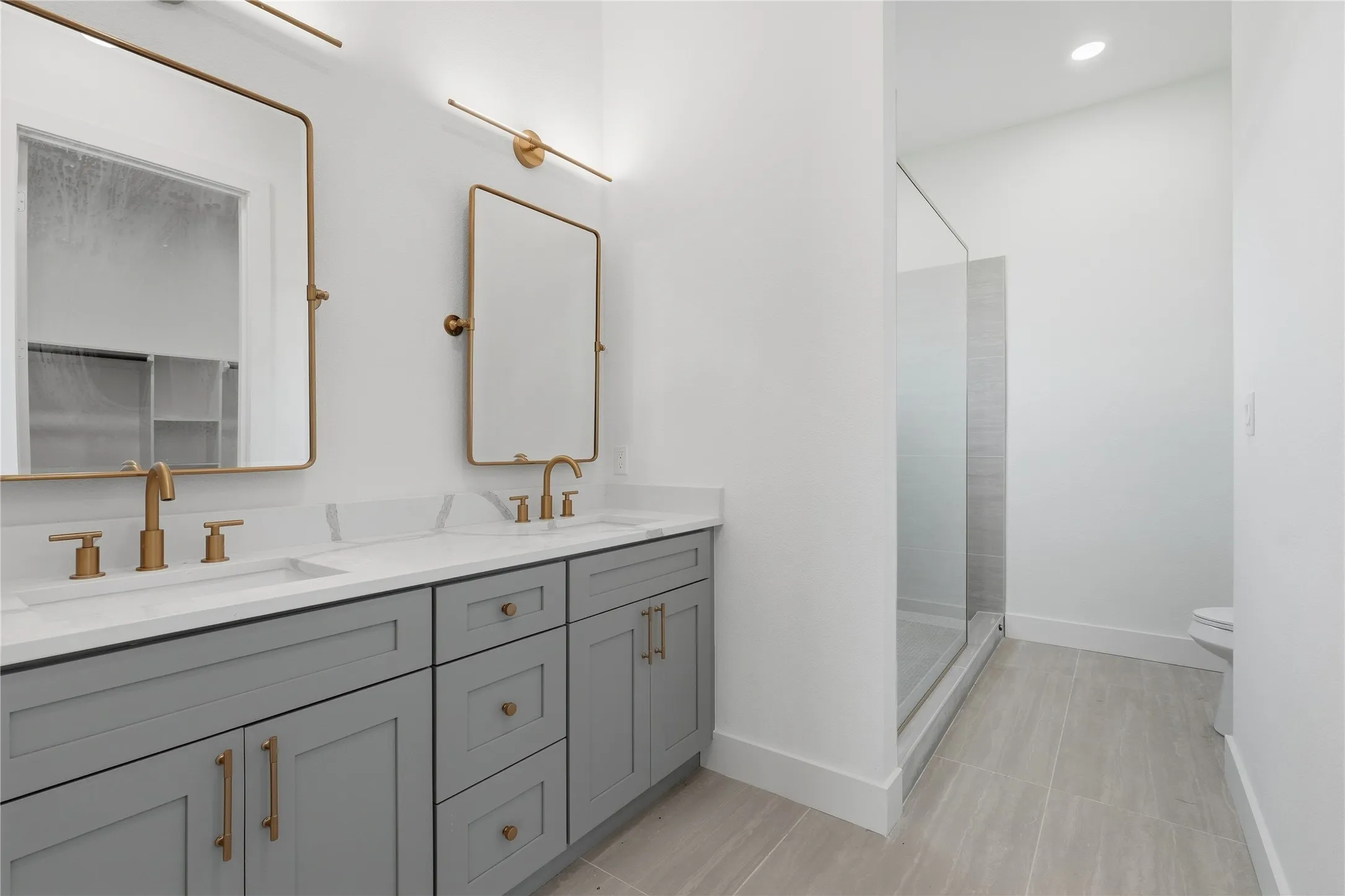 Dual vanity with gray cabinetry and gold-tone hardware, white countertops, two wall-mounted rectangular mirrors, and a glass-enclosed shower