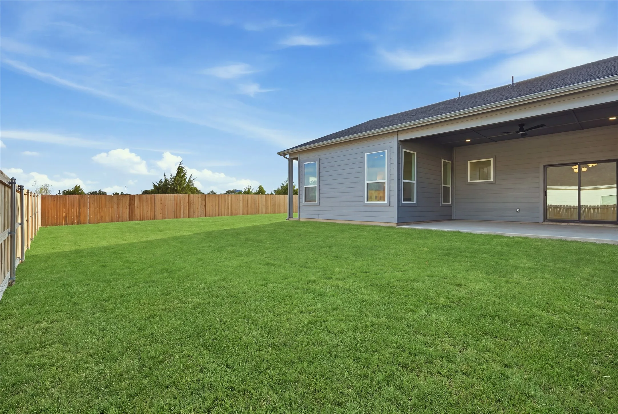 Expansive green lawn, wood privacy fencing, covered patio with integrated lighting, sliding glass door access, and multiple windows