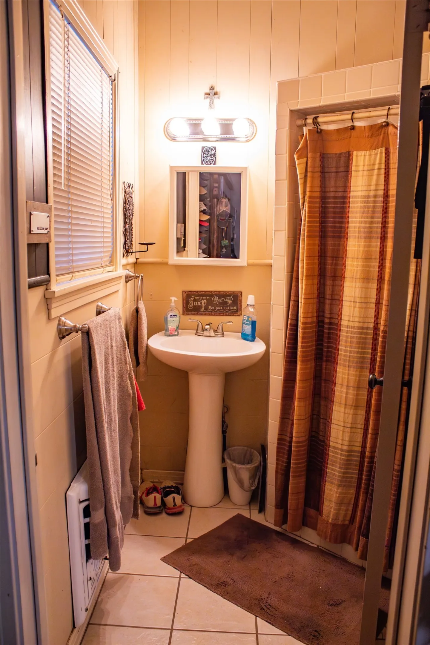 Pedestal sink, wall-mounted medicine cabinet, overhead vanity lighting, shower enclosure with tiled surround, and tile flooring