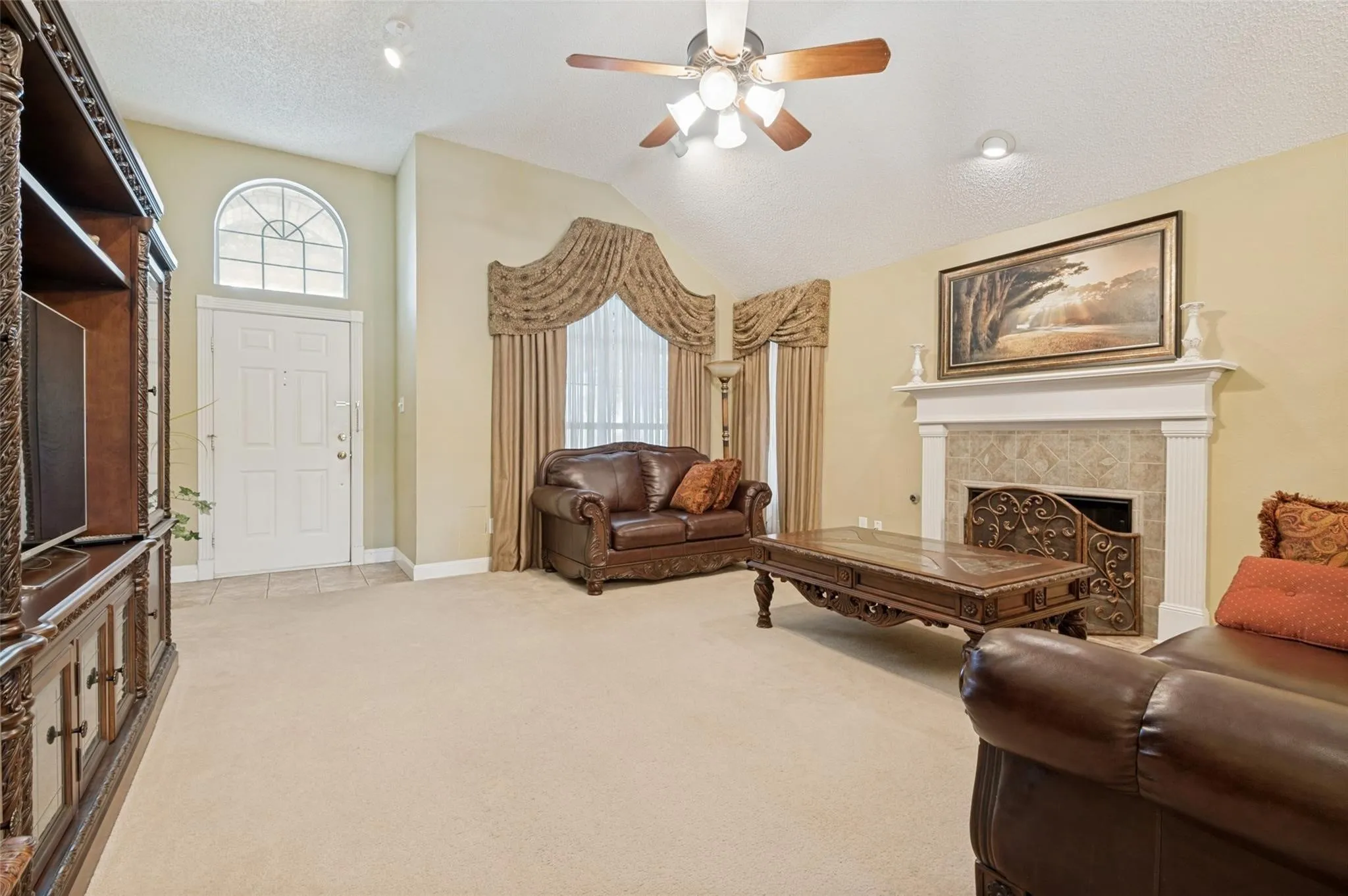 Vaulted ceiling with a ceiling fan and recessed lighting, white wood-frame fireplace with a stone surround, arched window above the entry door, and carpet flooring