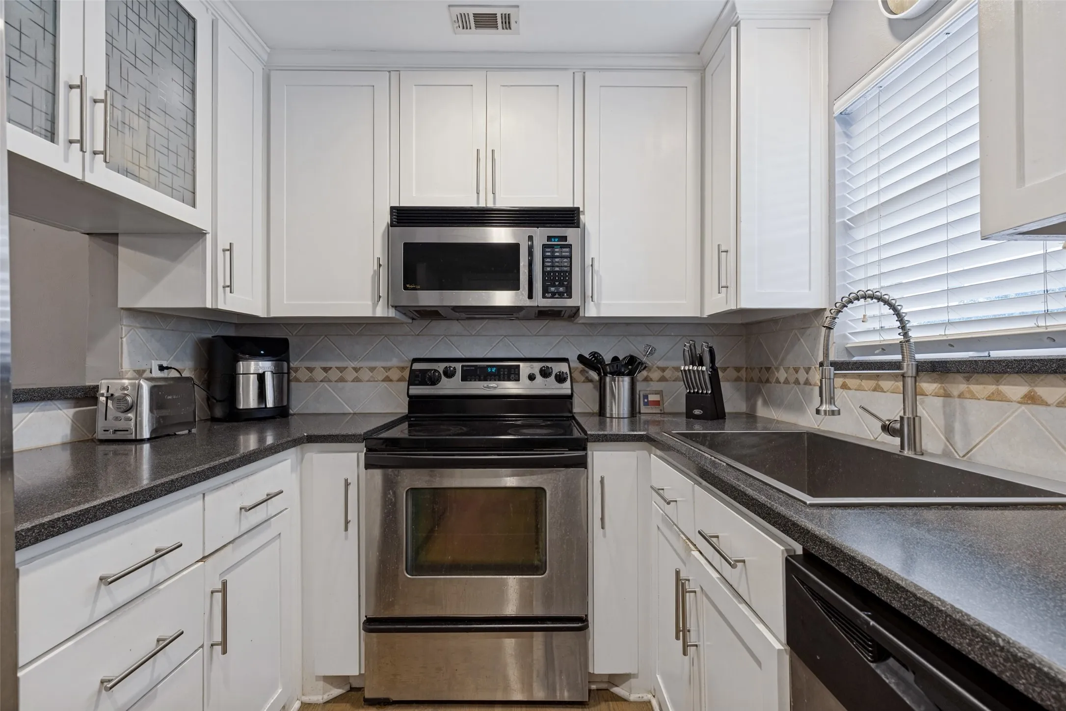 White cabinetry, dark countertops, stainless steel appliances, tile backsplash, and a stainless steel sink with a gooseneck faucet
