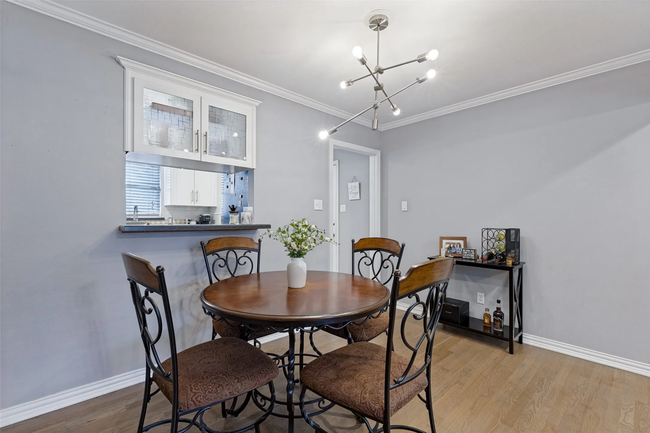 Wood-finish flooring, crown molding, modern linear chandelier, and a pass-through window with upper cabinetry
