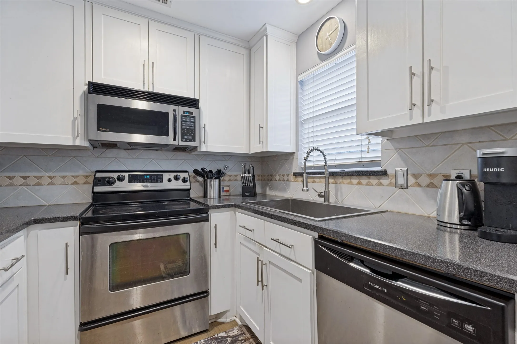 White shaker-style cabinetry, stainless steel appliances, dark countertops, tile backsplash, and a stainless steel sink