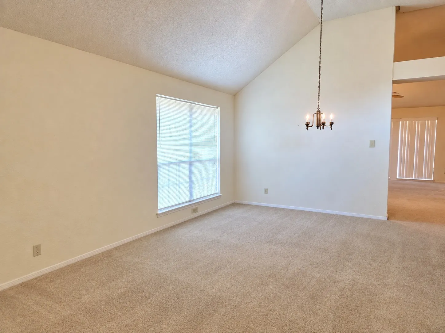 Vaulted ceiling with a suspended chandelier, large window with blinds, and neutral carpeting. VIRTUALLY DECLUTTERED USING AI
