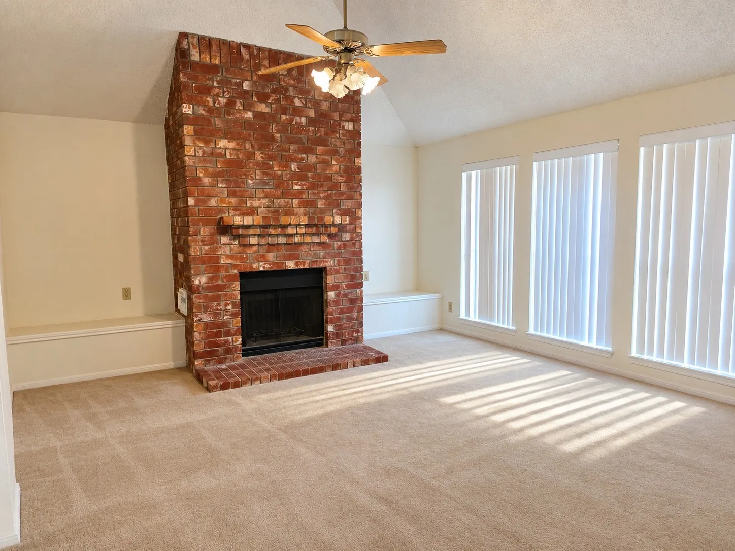 Vaulted ceiling with a ceiling fan, brick fireplace, built-in bench seating, multiple windows with vertical blinds, and carpeted flooring. VIRTUALLY DECLUTTERED USING AI