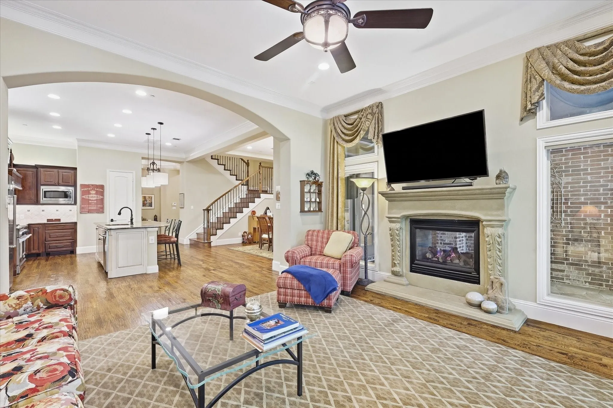 Hardwood flooring, arched entryway, ceiling fan, fireplace with stone surround, and built-in kitchen cabinetry