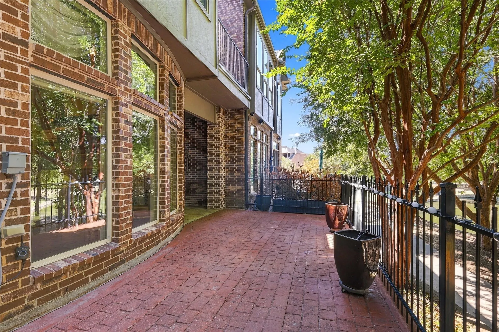 Curved brick exterior with multiple large windows, a red brick-patterned patio, black metal fencing, and mature trees