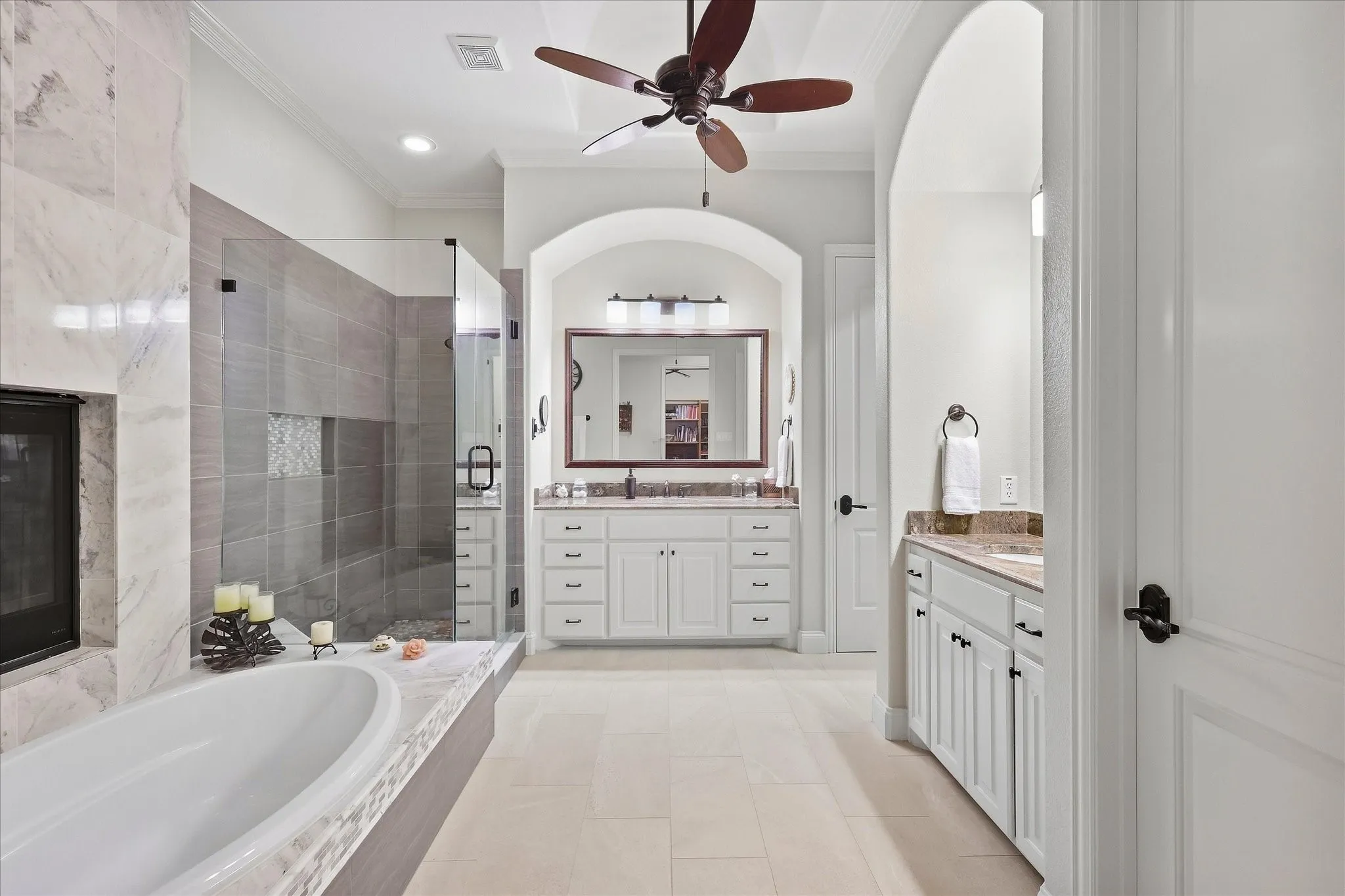 Freestanding soaking tub with tiled surround, glass-enclosed shower with tile work, dual vanities with stone countertops, and a ceiling fan