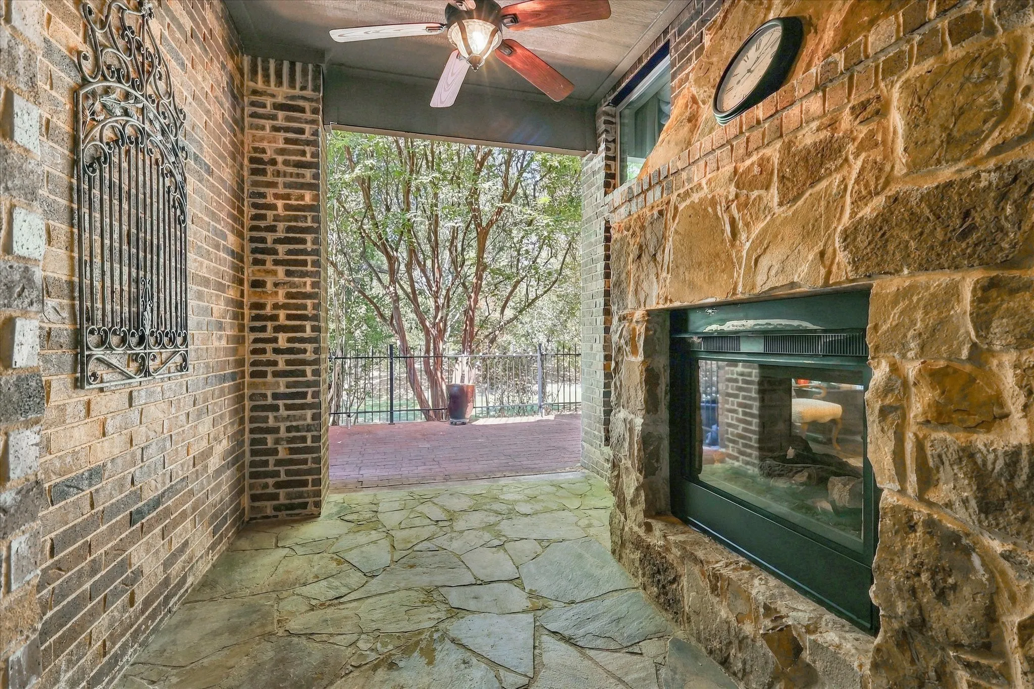Covered outdoor living space featuring flagstone flooring, brick walls, a stone fireplace, and a ceiling fan