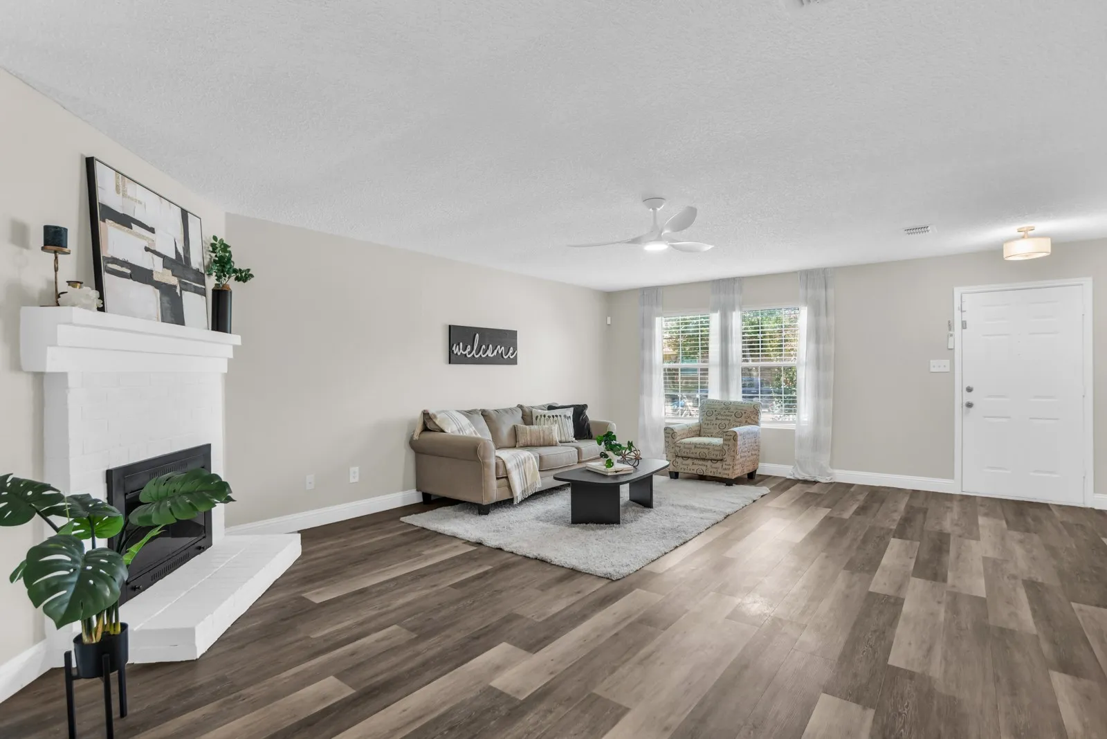 Spacious living area featuring wood-finish flooring, a white-painted fireplace with black firebox, window with blinds and sheer curtains, and a ceiling fan with integrated lighting
