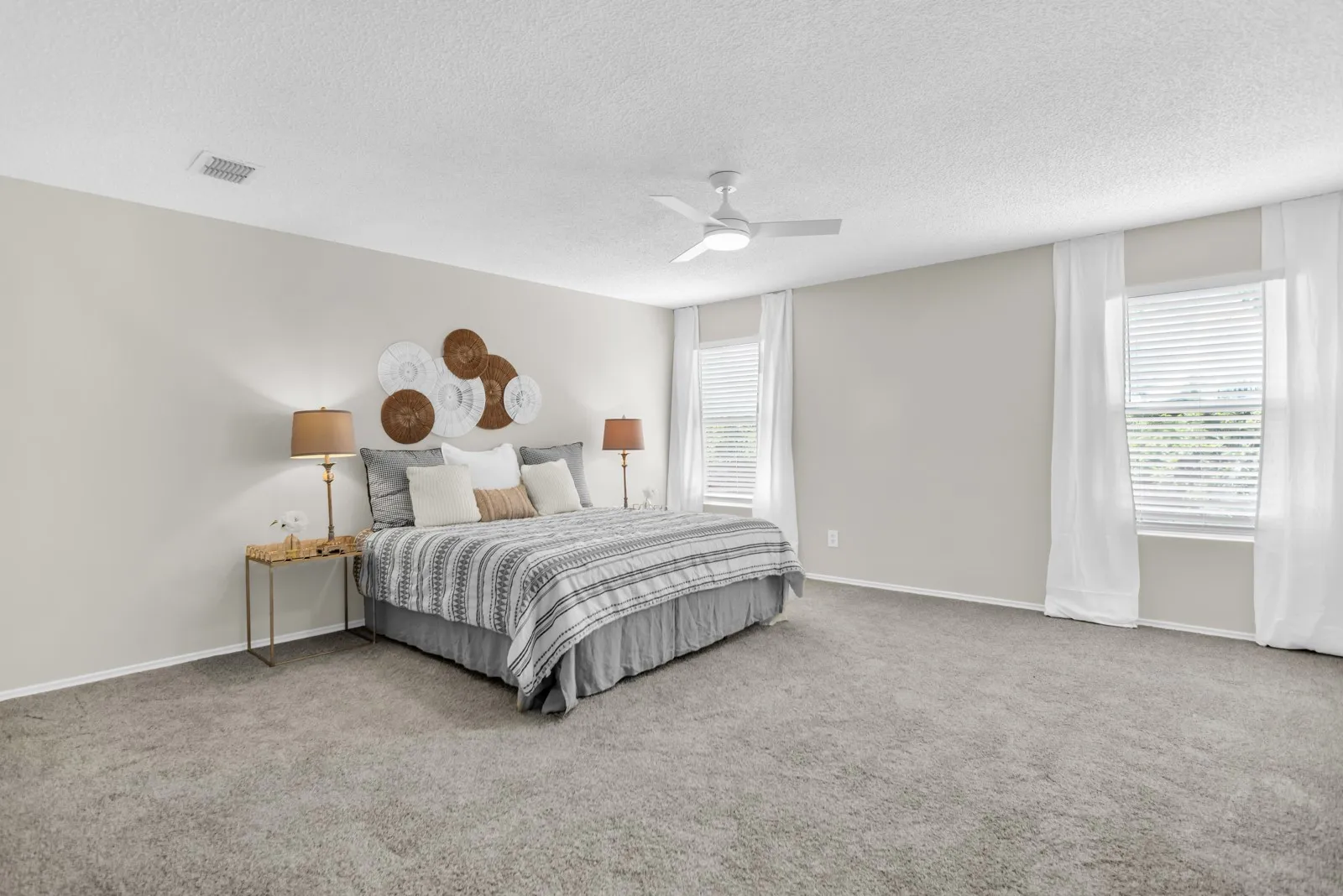 Spacious room with plush carpeting, neutral wall tones, two large windows with blinds, and a ceiling fan