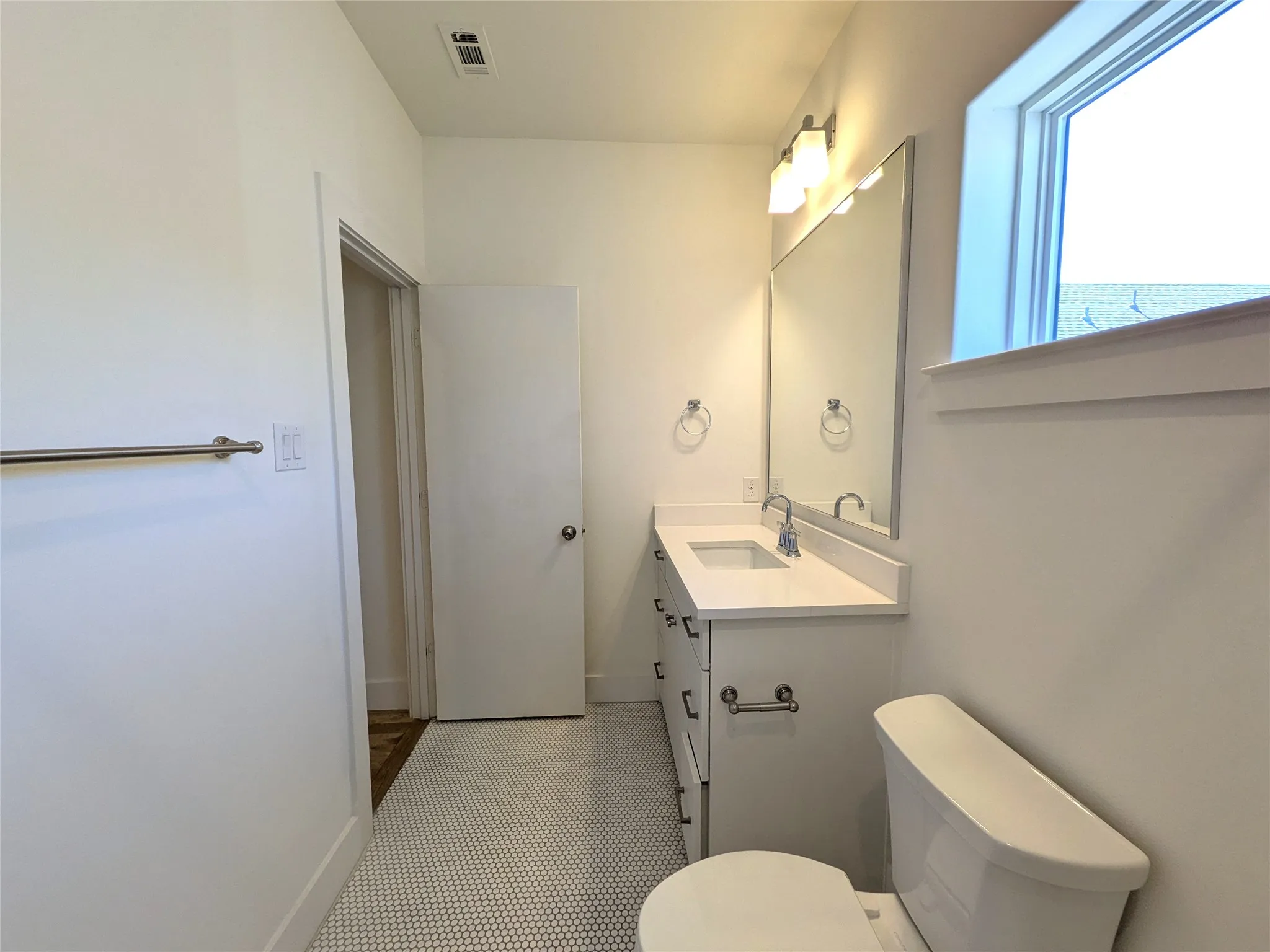 White penny tile flooring, a white vanity with an integrated sink, a large wall mirror, and a window for natural illumination