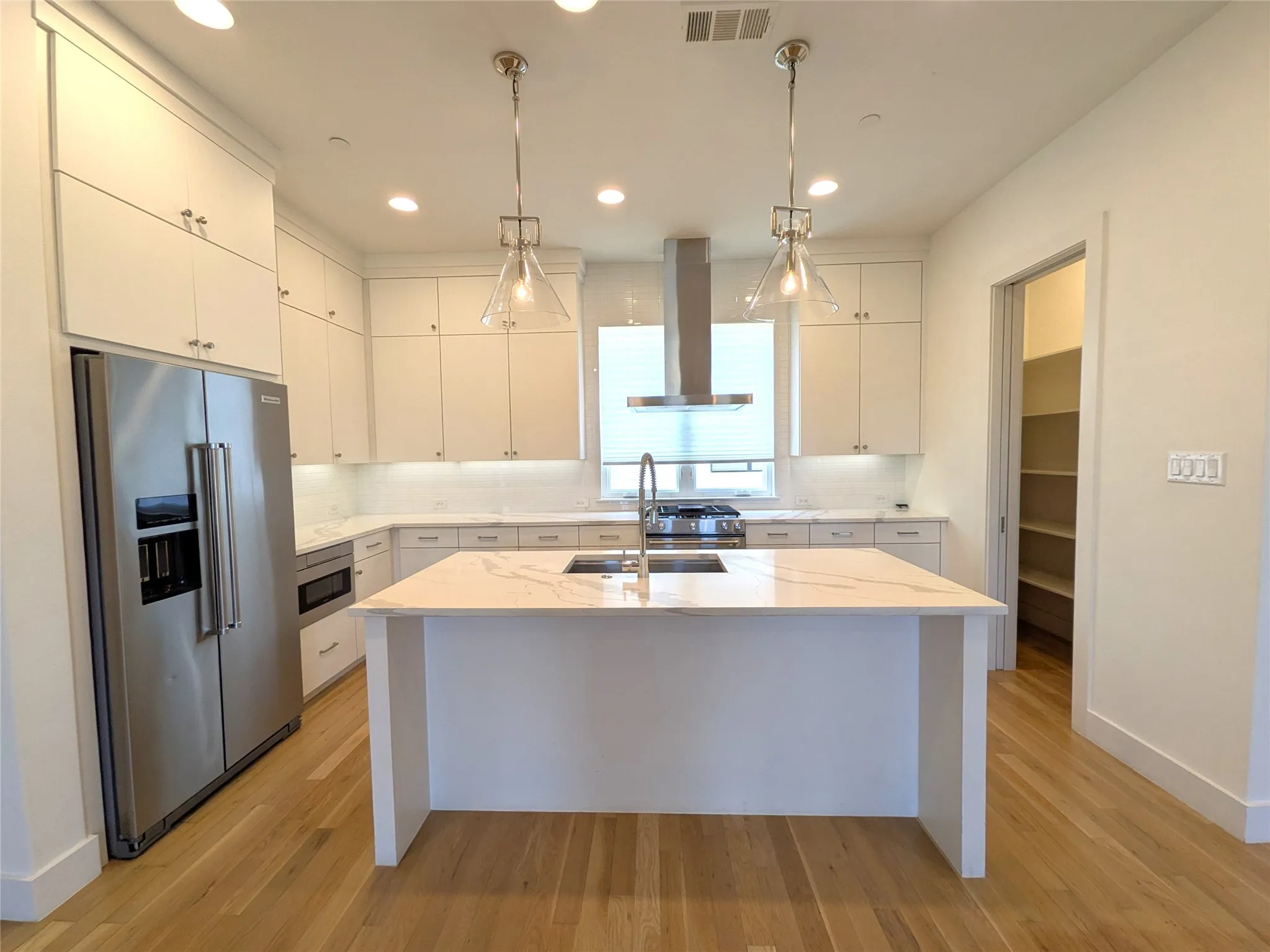 Modern kitchen featuring a large central island with an integrated sink, stainless steel appliances, white flat-panel cabinetry, wood-finish flooring, and a walk-in pantry