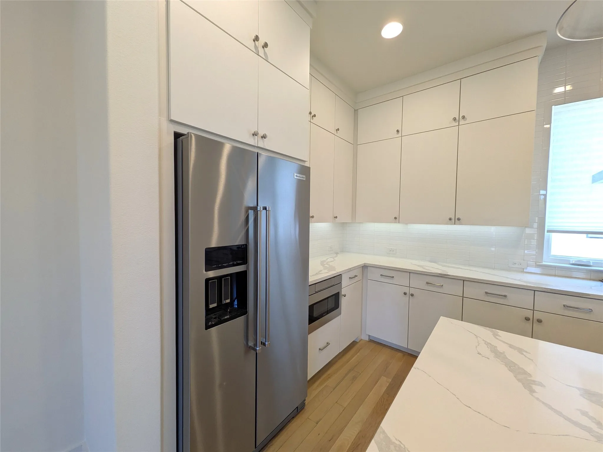Stainless steel refrigerator, white shaker-style cabinetry, light wood-finish flooring, white subway tile backsplash, and stone countertops