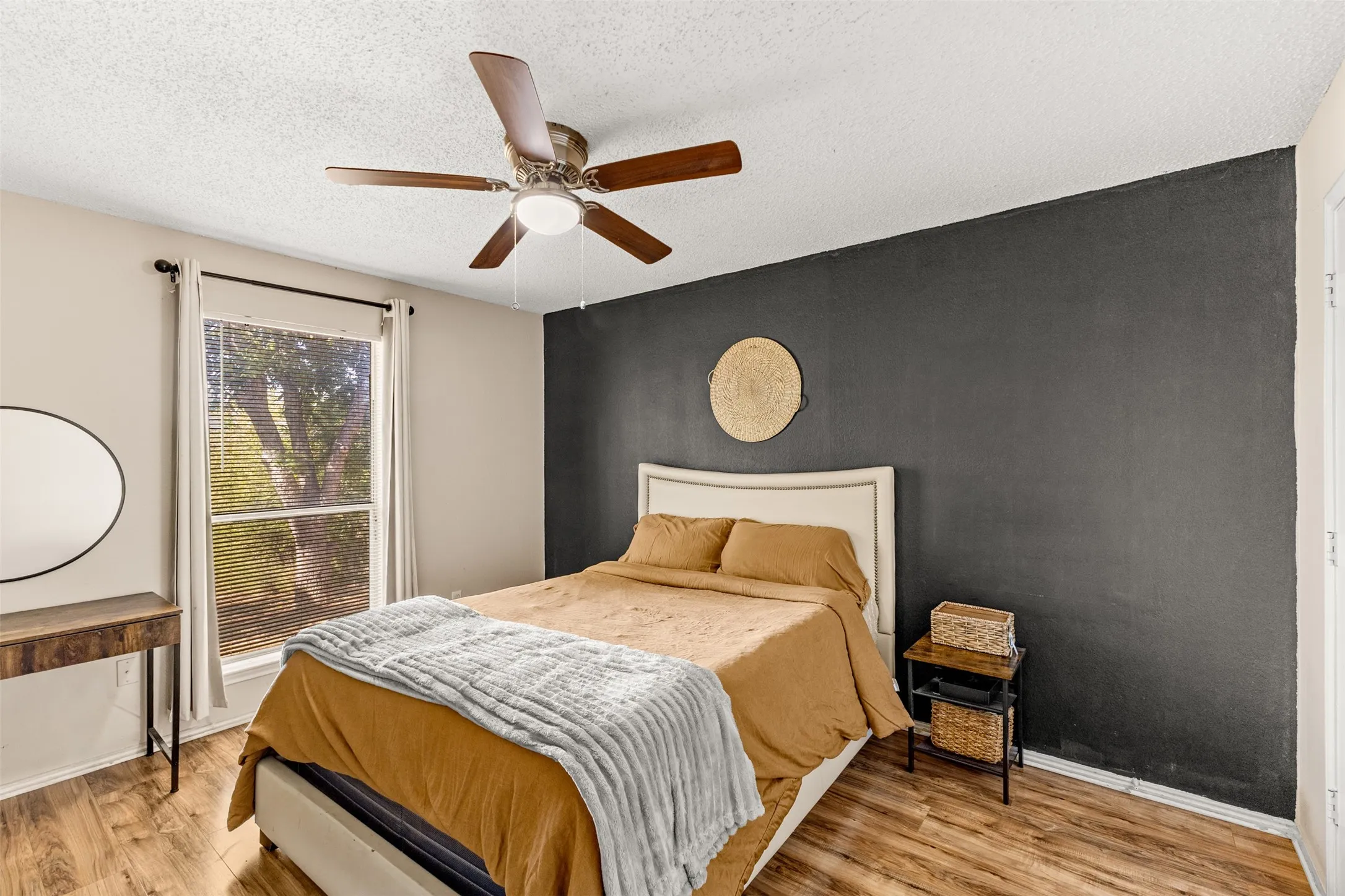 Wood-finish flooring, ceiling fan with integrated lighting, window with blinds, dark accent wall, and a headboard with upholstered details