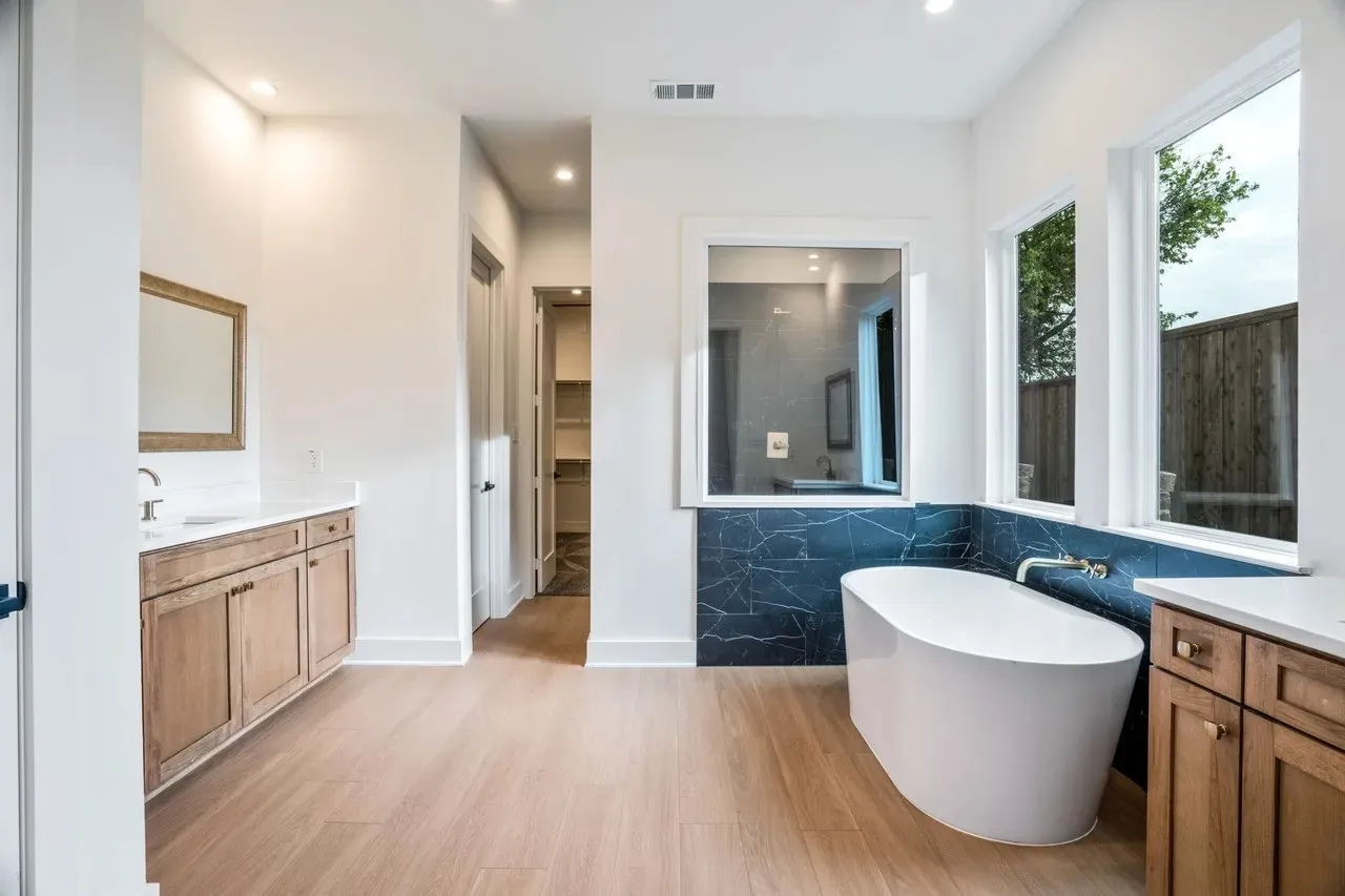 Freestanding soaking tub with dark-toned accent tile surround, wood-finish flooring, dual vanity with light-toned countertops, and recessed lighting
