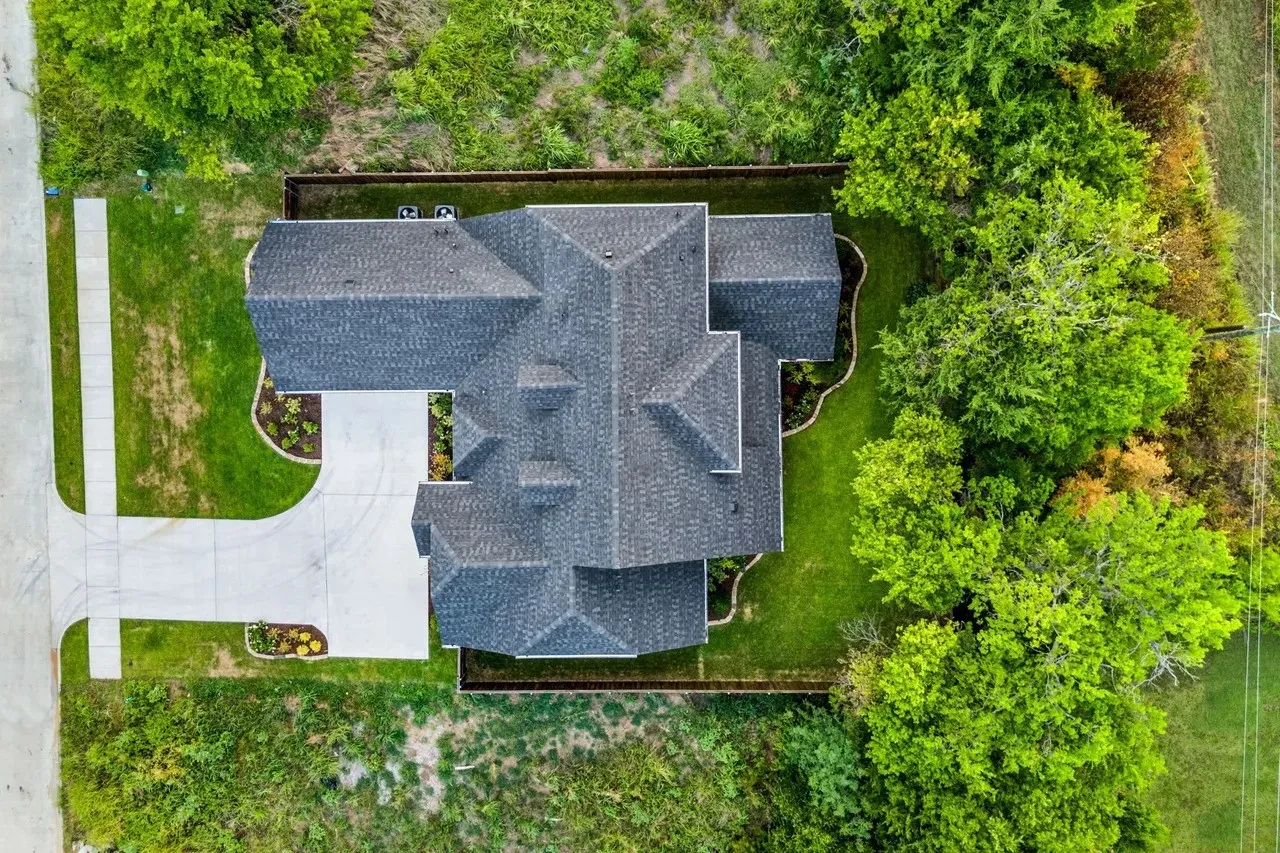 Shingled roof with multiple gables, extensive concrete driveway, manicured lawn, established landscaping, and mature perimeter trees