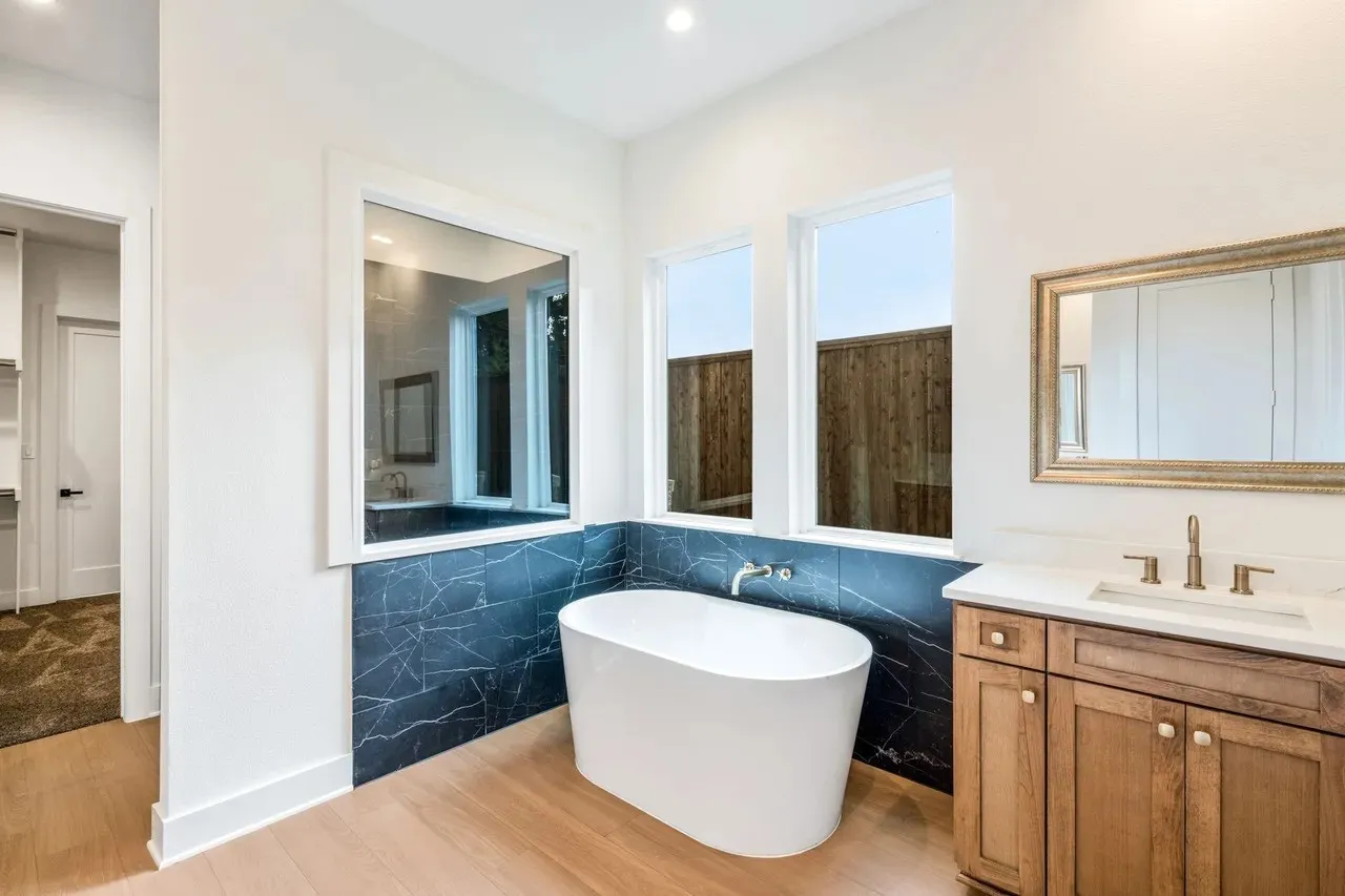 Freestanding soaking tub, floor-mounted tub filler, dark tiled accent wall, single vanity with wood cabinetry, and a framed vanity mirror