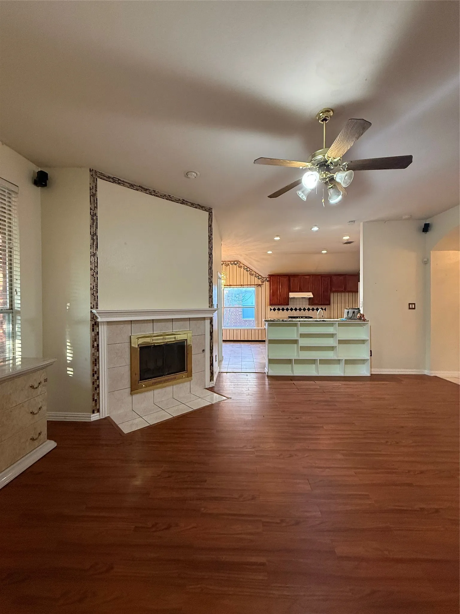 Wood-finish flooring throughout, featuring a tiled fireplace with a gold-tone insert, a ceiling fan, and built-in shelving