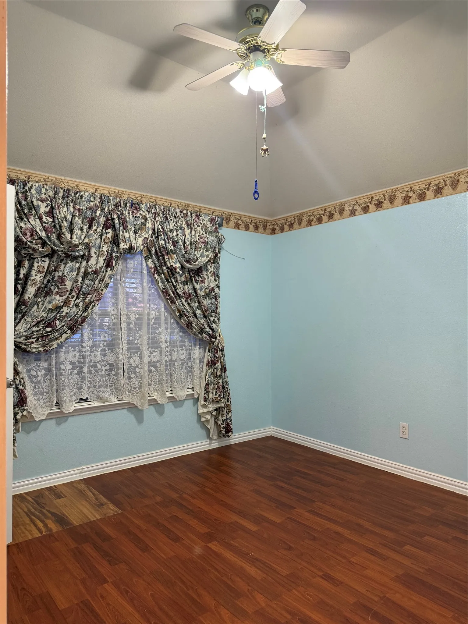 Wood-finish flooring, ceiling fan with light fixture, window with drapes, and light blue walls