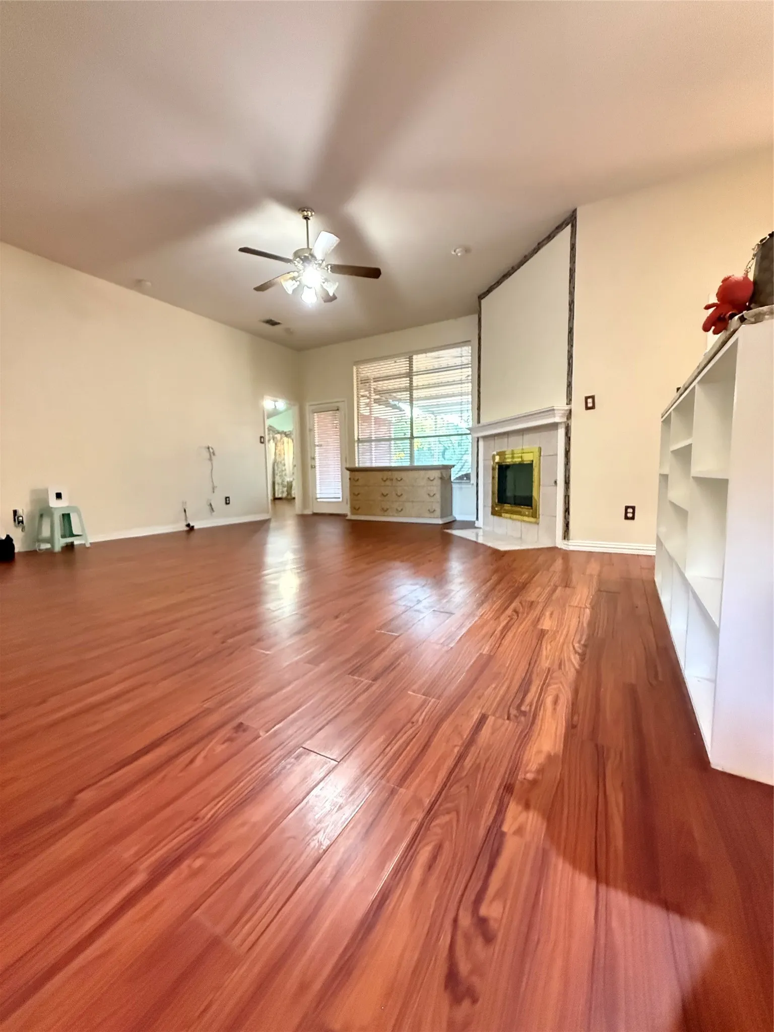 Wood-finish flooring throughout, vaulted ceiling with a ceiling fan, a corner fireplace with a mantel, built-in shelving, and a glass-paneled door