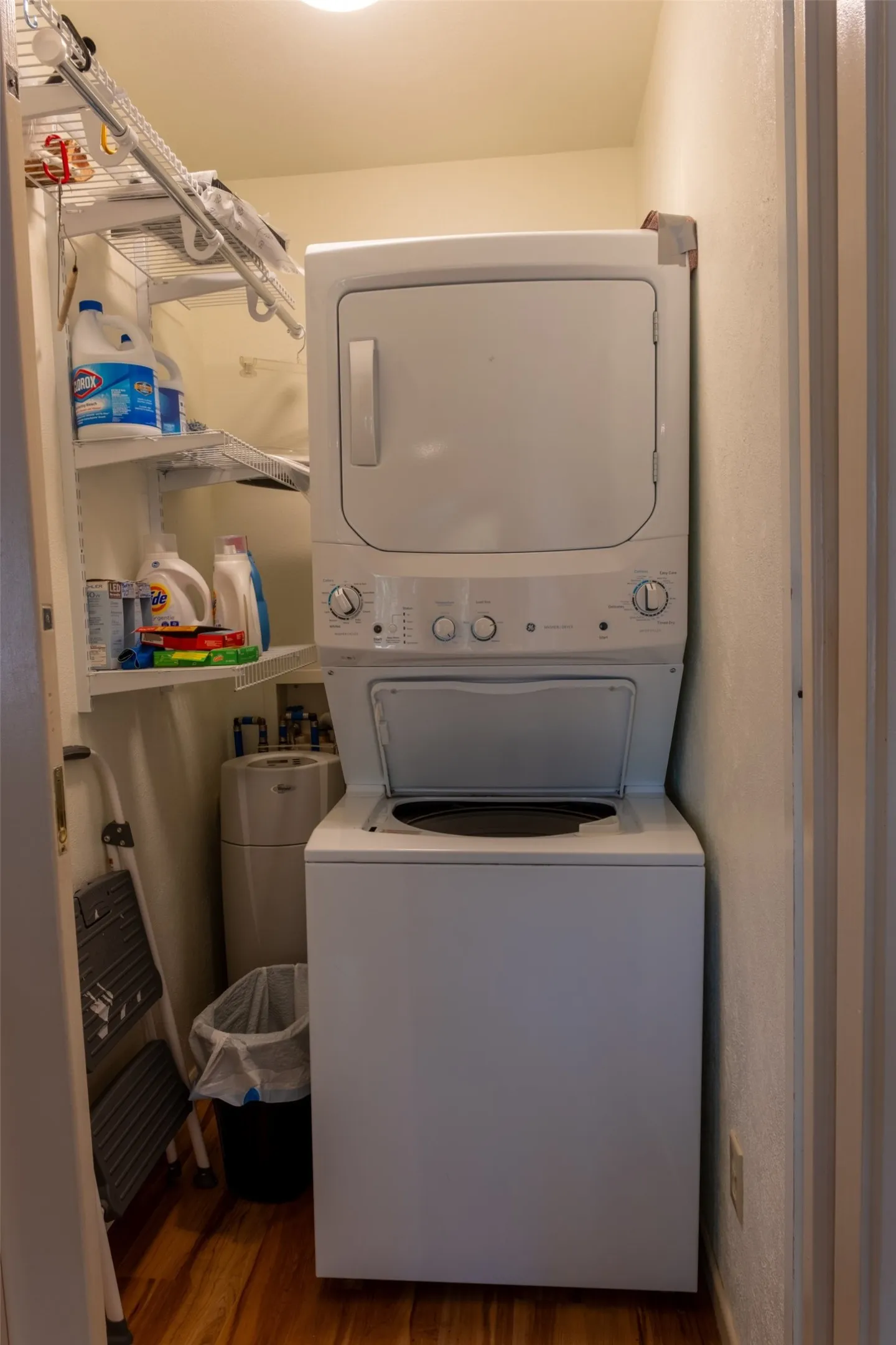 Compact laundry area featuring a stacked washer and dryer unit, wood-finish flooring, and built-in wire shelving