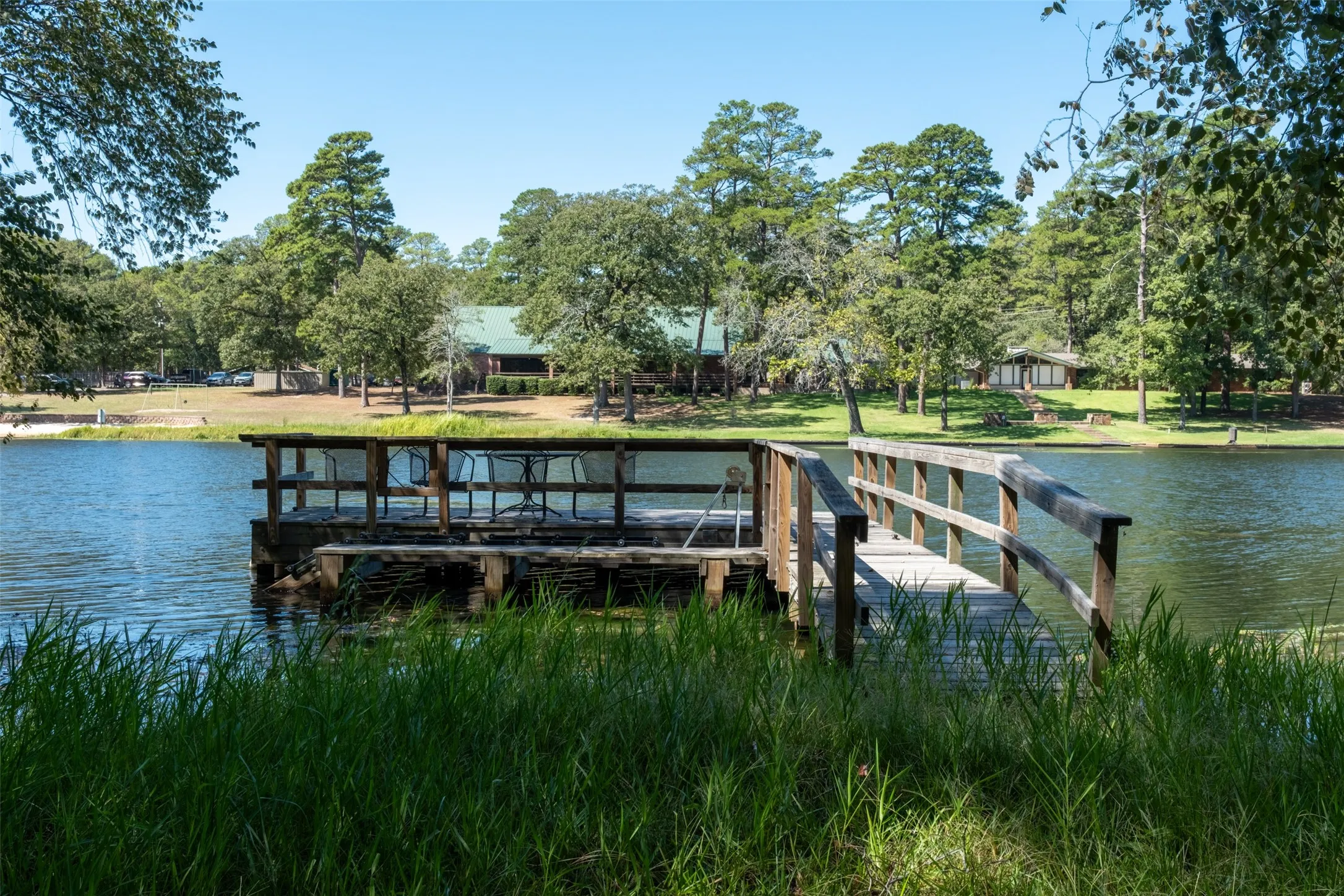 Wood-plank dock with a built-in seating area over a body of water, surrounded by mature trees and a distant building with a green roof