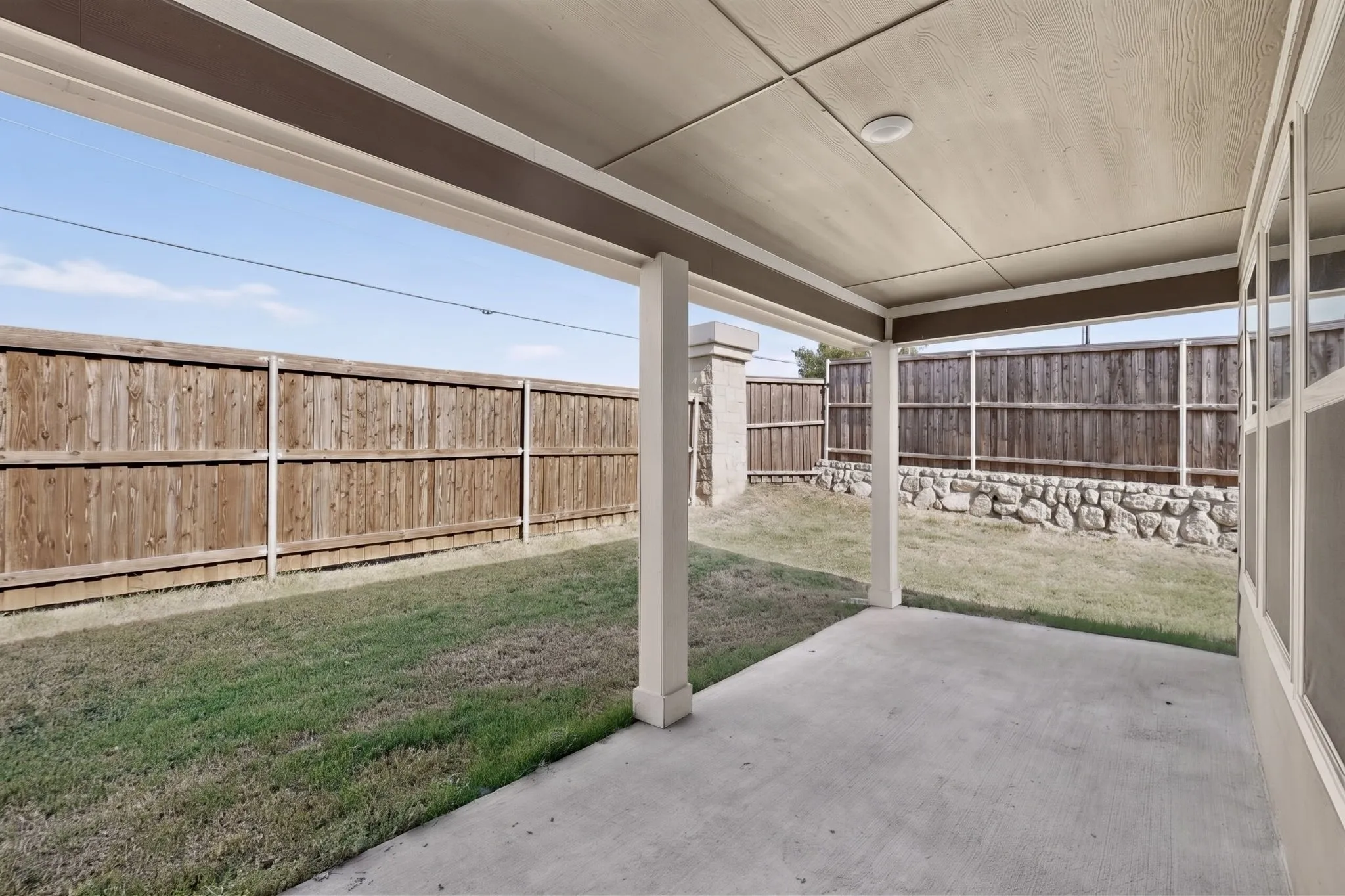 Covered concrete patio with recessed lighting, bordered by a wood privacy fence and a low stone wall