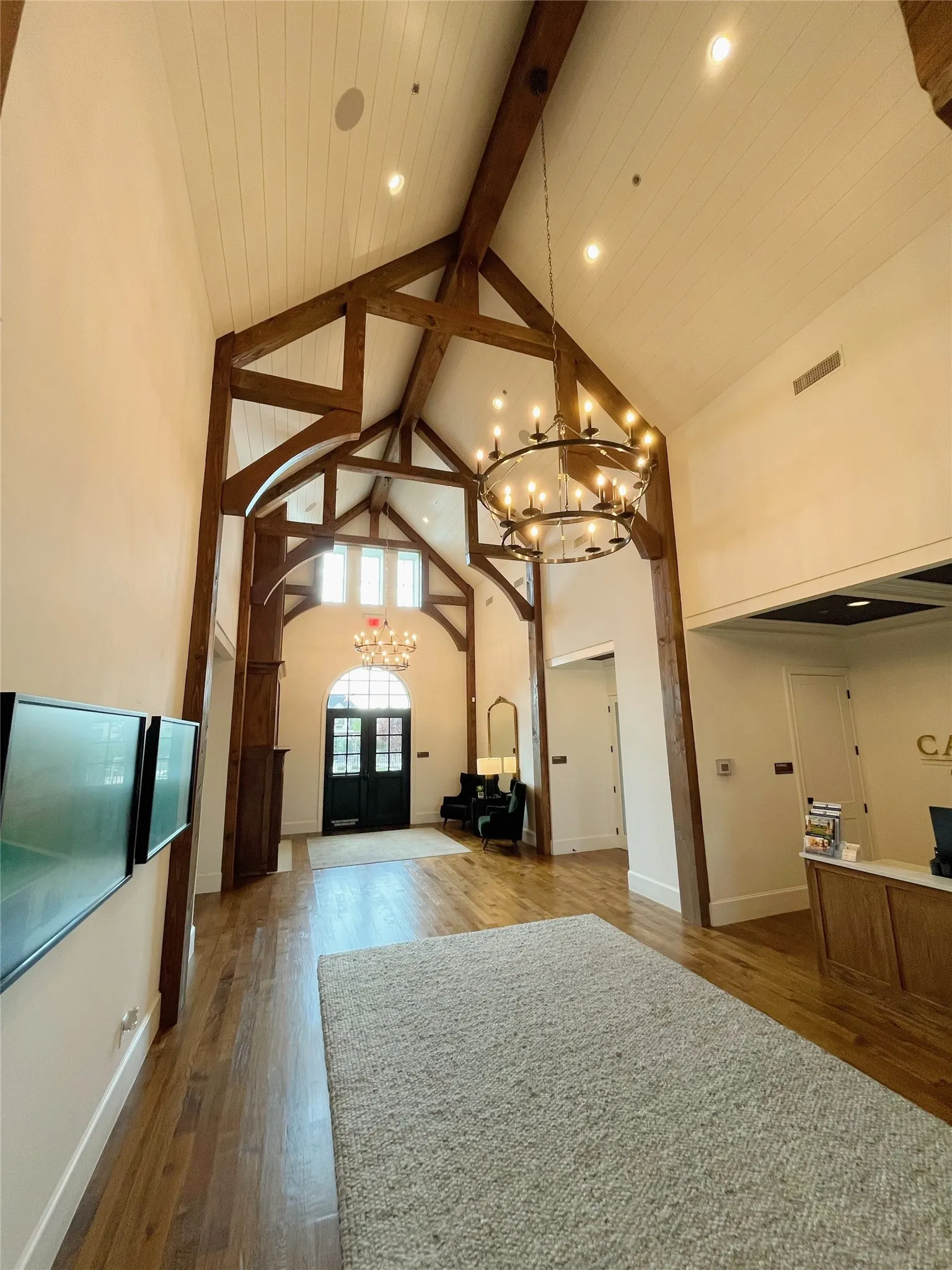 Vaulted ceilings with exposed wood beams, wood-finish flooring, two large chandeliers, and a prominent arched entryway