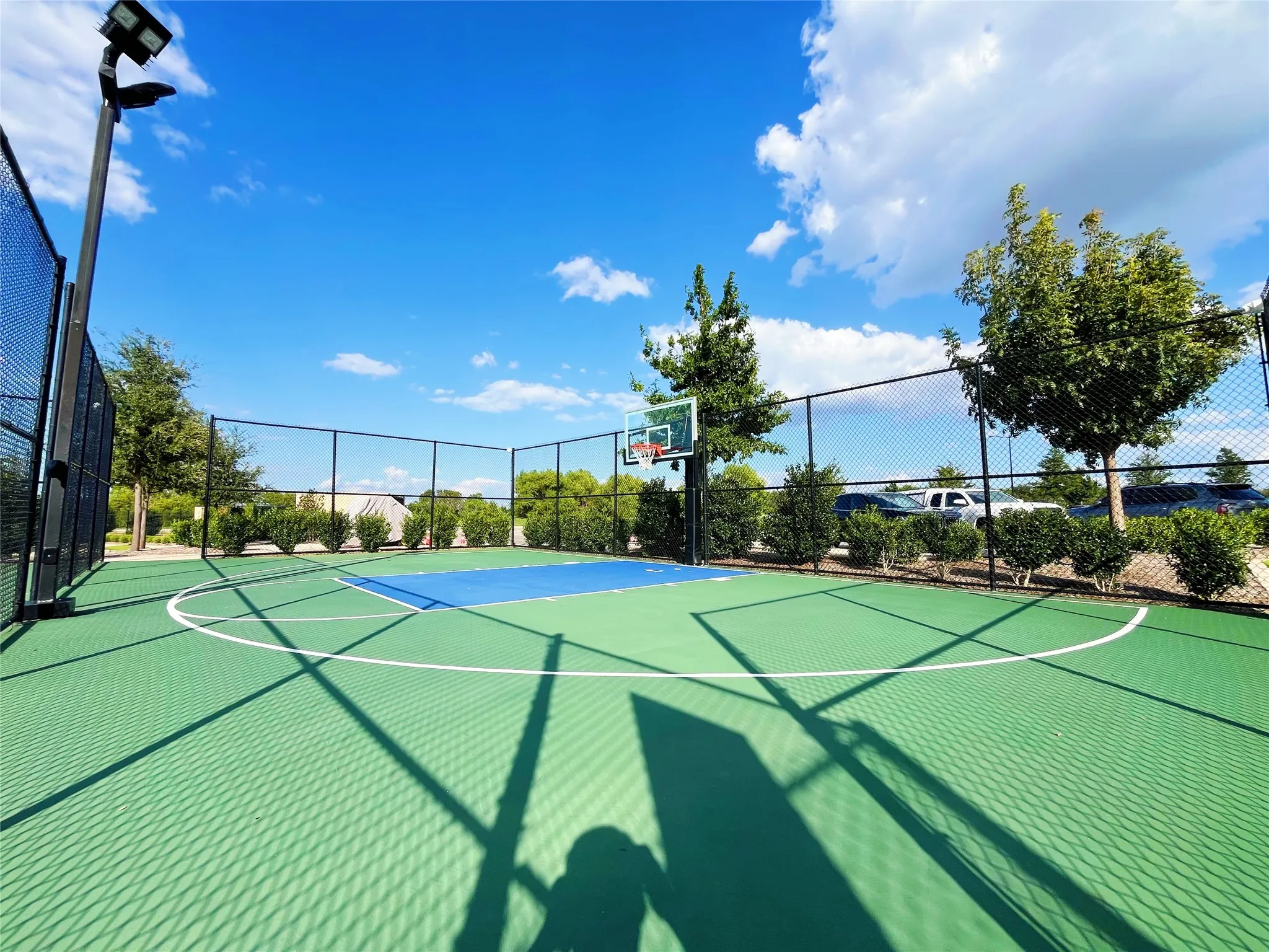Fenced multi-sport court with vibrant green and blue surfacing, equipped with a basketball hoop