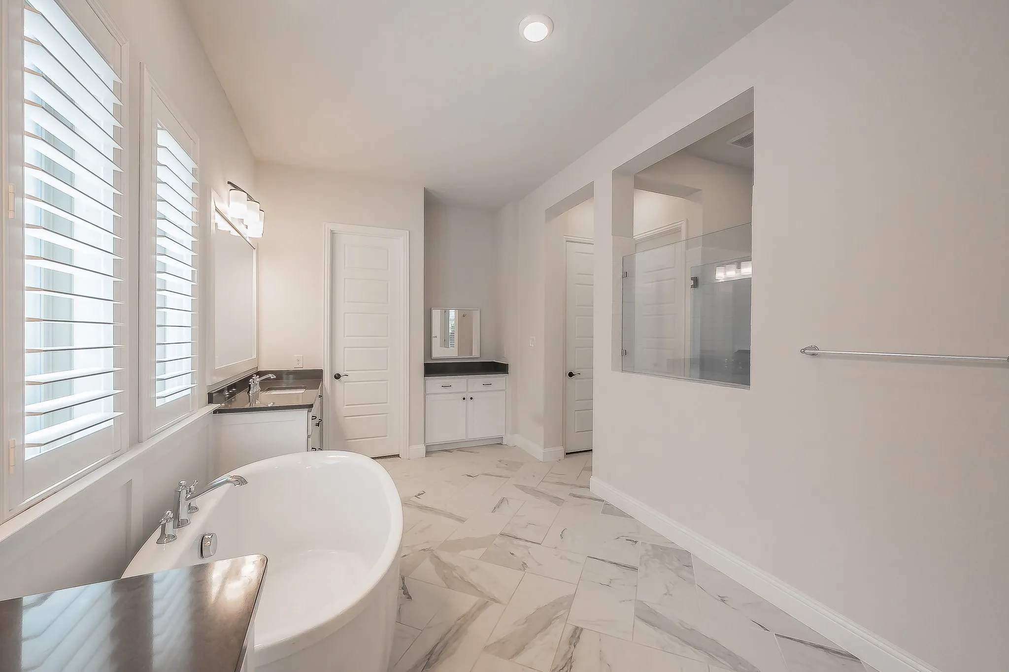 Freestanding soaking tub, dual vanity with dark countertops, white plantation shutters, glass-enclosed shower, and large format tile flooring