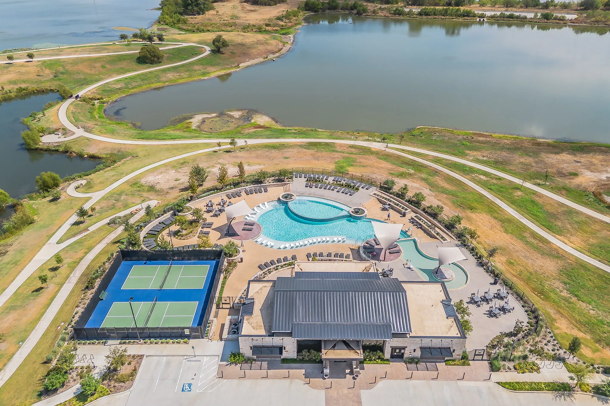 Expansive outdoor pool area with surrounding lounge seating, two tennis courts, community building with metal roof, and multiple winding pedestrian paths bordering a body of water