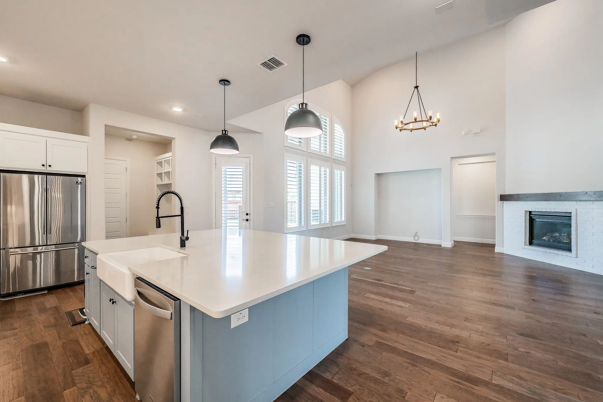 Open-concept interior featuring wood-finish flooring, a kitchen island with a farmhouse sink, stainless steel appliances, and a fireplace with a decorative mantel