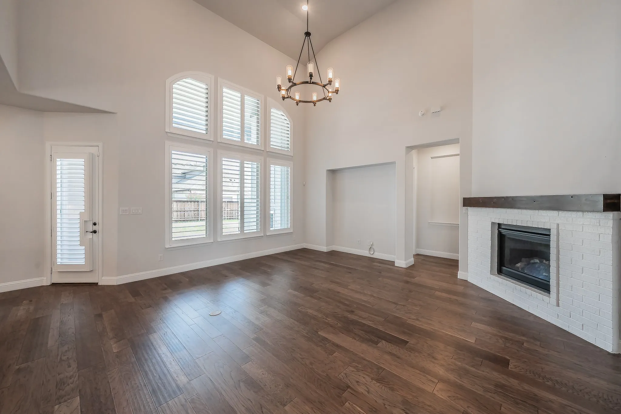 Vaulted ceiling with an overhead chandelier, wood-finish flooring, a contemporary fireplace with a white brick surround and wood mantle, and multiple windows featuring plantation shutters