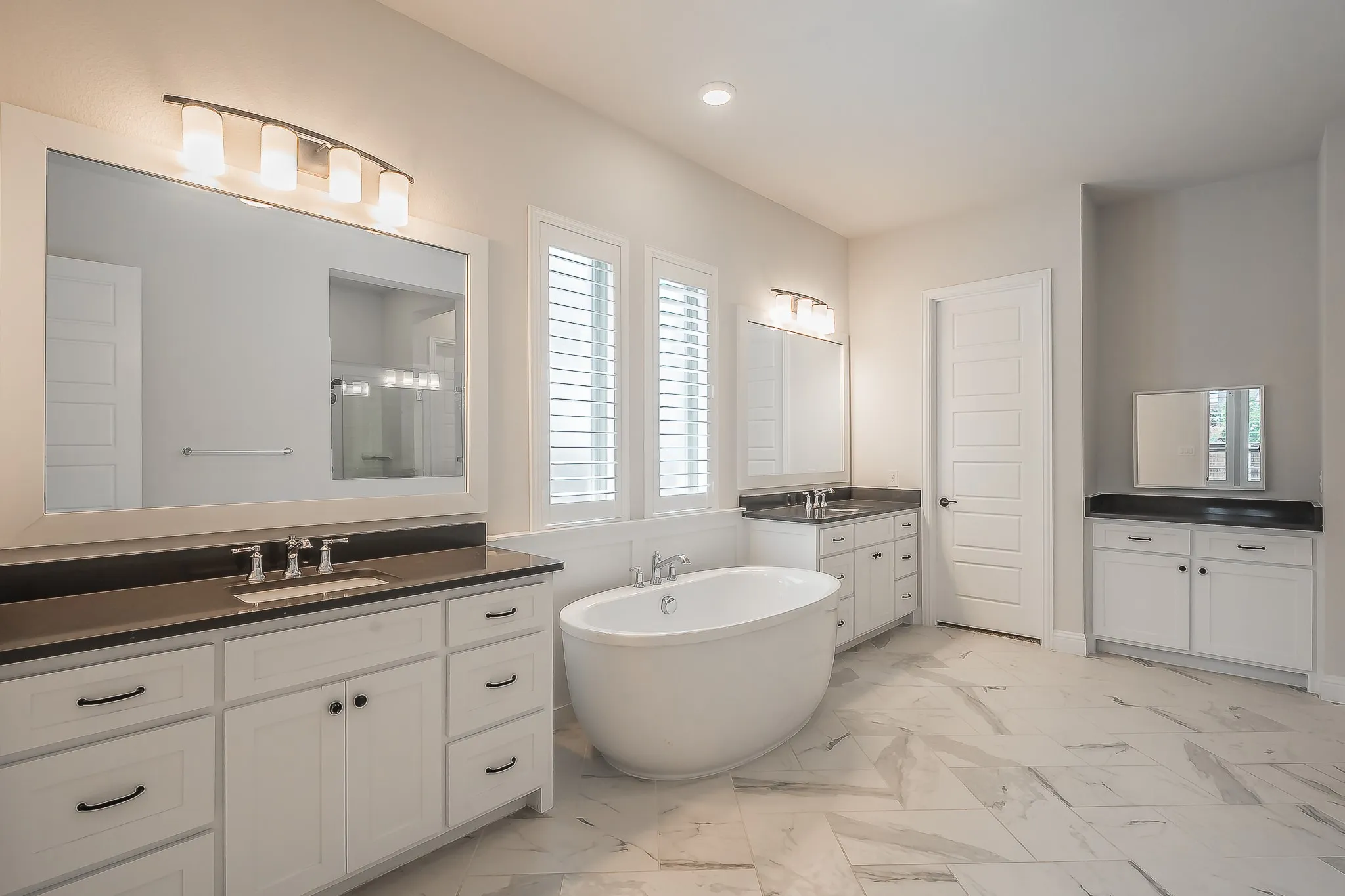 Freestanding soaking tub, dual vanities with dark countertops, white cabinetry with dark hardware, shuttered windows, and large format floor tiling