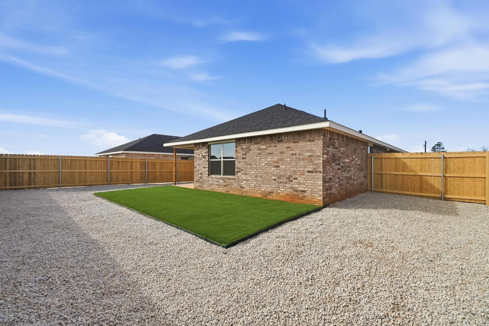 Exterior featuring brick siding, a dark shingle roof, a privacy fence, and a gravel yard with a dedicated artificial turf area